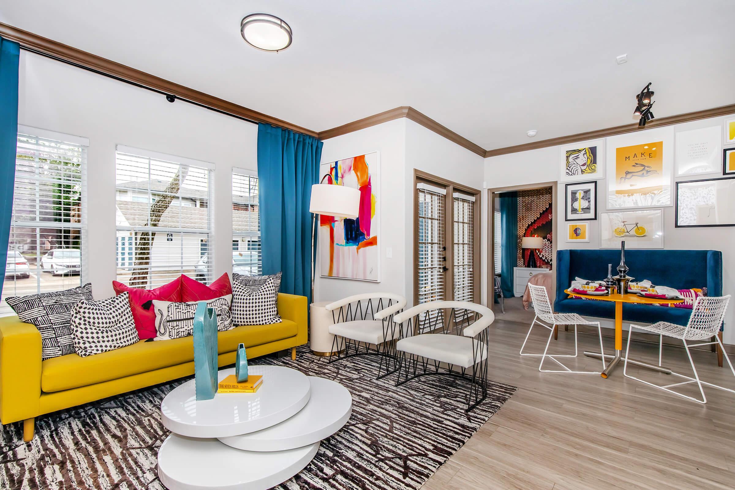 A bright living room featuring a yellow sofa adorned with colorful pillows, accompanied by a round white coffee table. Large windows with blue curtains allow natural light in. A blue dining area is visible in the background, decorated with art and a vibrant atmosphere. The floor is covered with a stylish area rug.