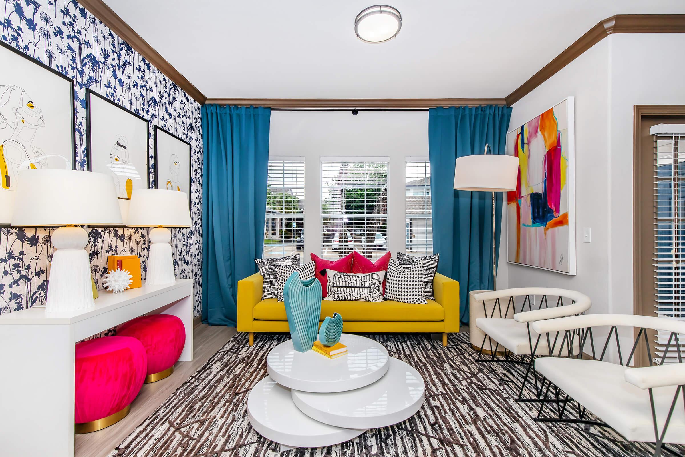 A bright and stylish living room featuring a yellow sofa with colorful throw pillows, two pink accent chairs, a white coffee table, and decorative vases. The walls are adorned with patterned wallpaper and framed art. Large curtains in teal frame the windows, allowing natural light to brighten the space.