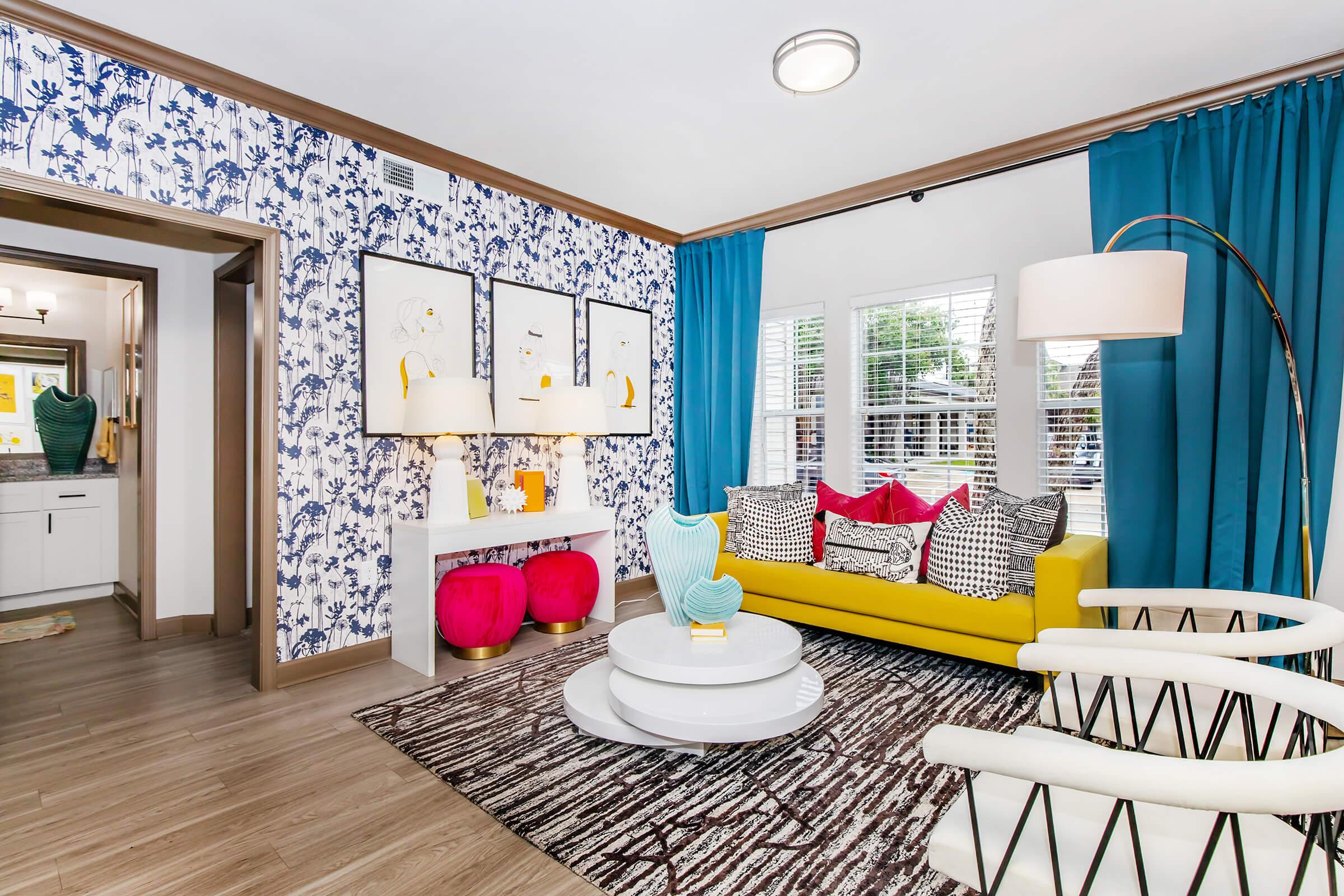 A vibrant living room featuring a yellow sofa adorned with colorful pillows, a round white coffee table, and stylish armchairs. The walls are decorated with blue floral wallpaper and matching curtains. A console table with decorative lamps and artwork adds charm to the space, which has warm wooden flooring.
