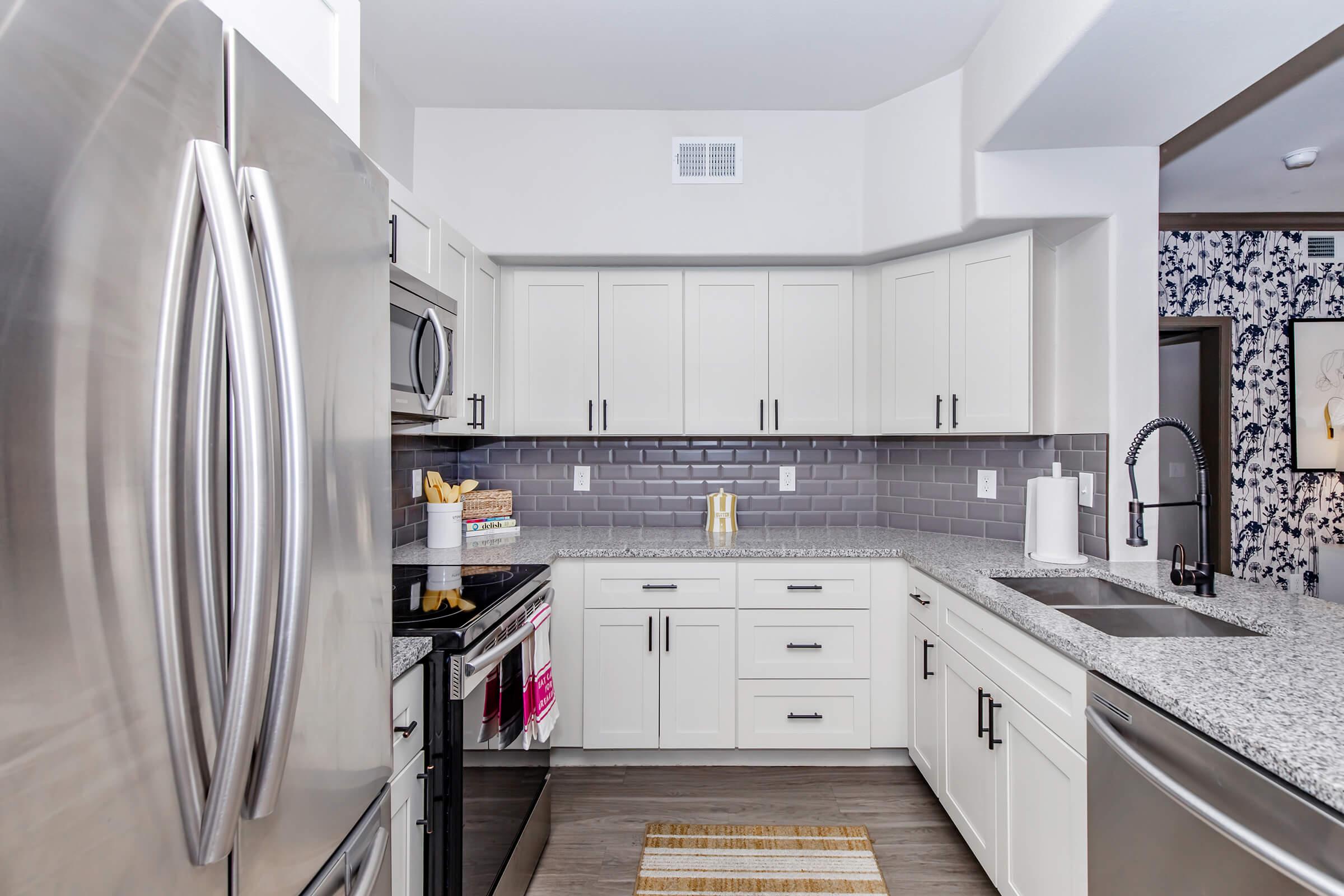 Modern kitchen featuring stainless steel appliances, a gray backsplash, and white cabinetry. The countertops are gray speckled stone, with utensils neatly arranged on one side. A patterned wall adds visual interest, and a small rug lies on the floor, enhancing the cozy atmosphere.
