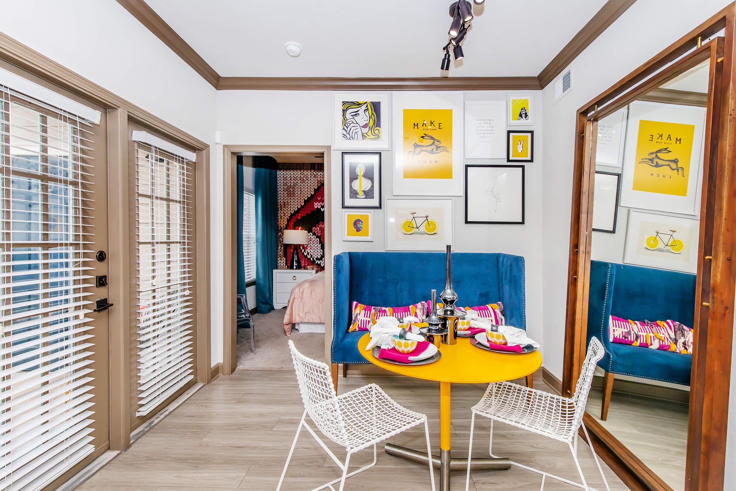 A bright and modern dining area with a round yellow table set for two. White wire chairs surround the table, which is adorned with colorful dishes. A large mirror reflects a blue sofa and vibrant artwork on the walls. Sunlight streams in through blinds, creating a warm and inviting atmosphere.