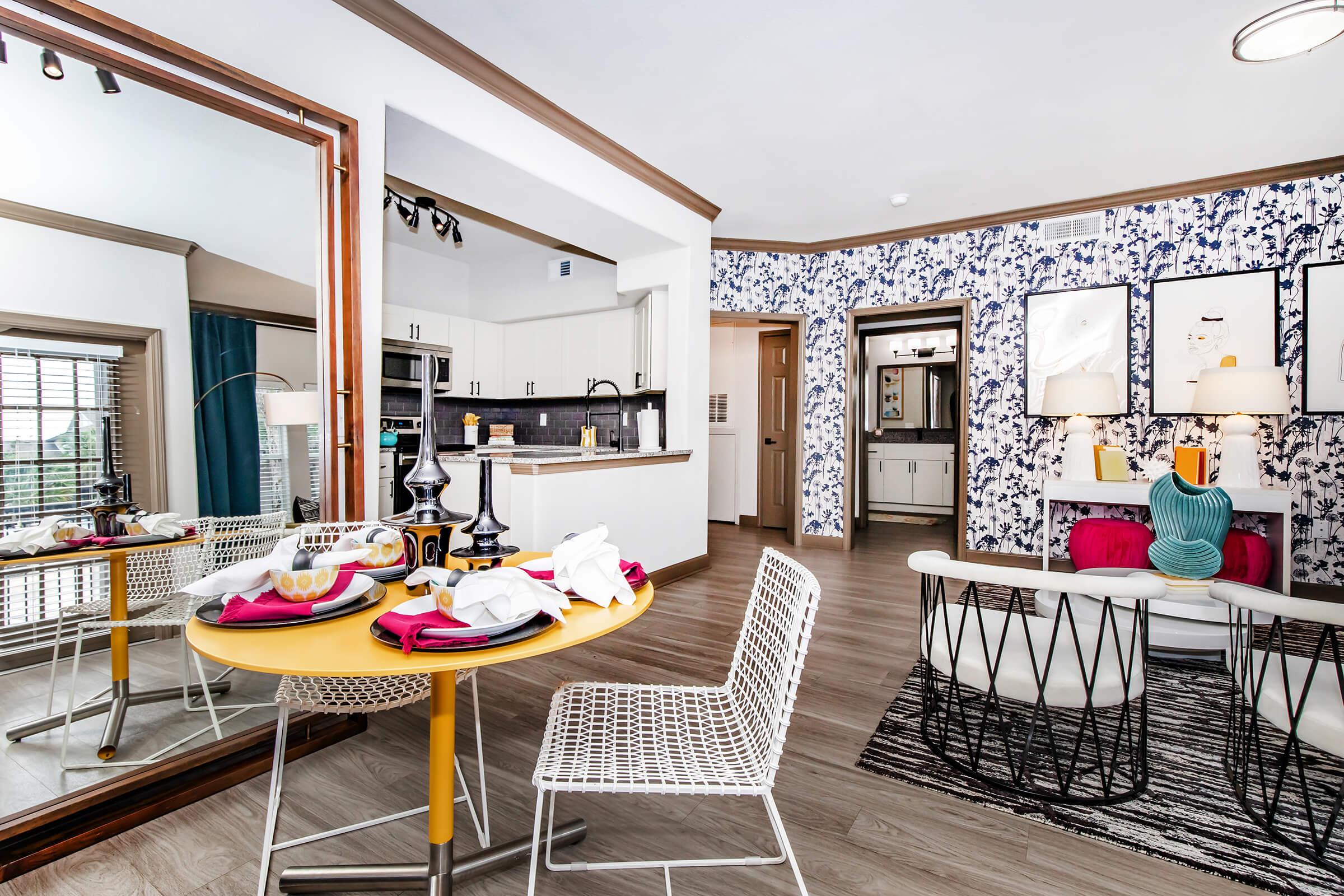 A modern dining area featuring a round yellow table set with dishes and napkins. Stylish white chairs surround the table, and a large mirror reflects the room. The decor includes a blue floral wallpaper, dark cabinetry, and a couple of colorful artworks on the wall. Natural light fills the space through windows.