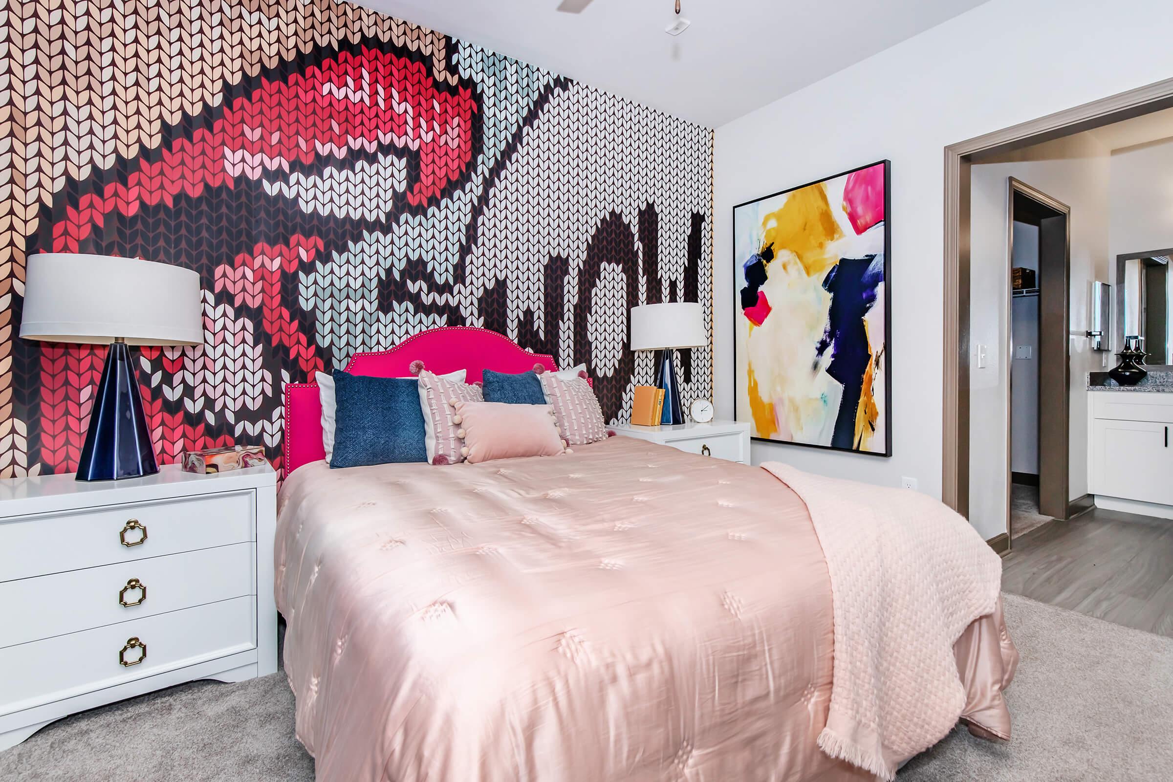 A modern bedroom featuring a large pink bedspread on a neatly made bed with decorative pillows. The wall behind the bed has a vibrant graphic mural, while a colorful abstract painting hangs nearby. Two stylish lamps sit on white nightstands, and an open doorway leads to a bathroom area.