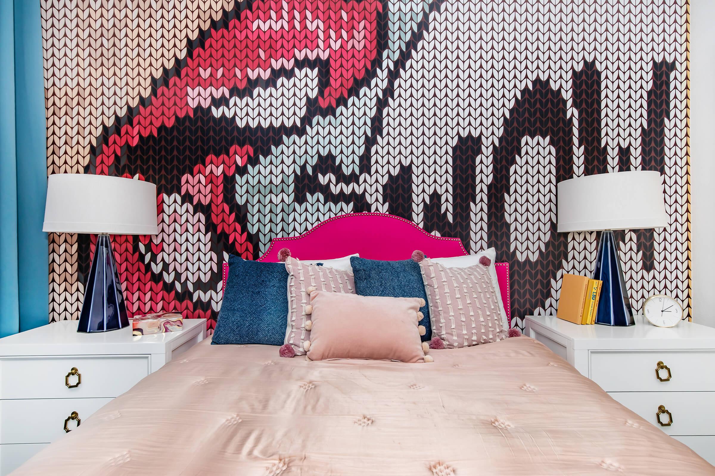 A stylish bedroom featuring a pink headboard with decorative pillows on a bed, flanked by two modern bedside tables with lamps. The wall behind the bed displays a large, colorful graphic design. A clock and a book are visible on the bedside tables, adding to the contemporary decor.