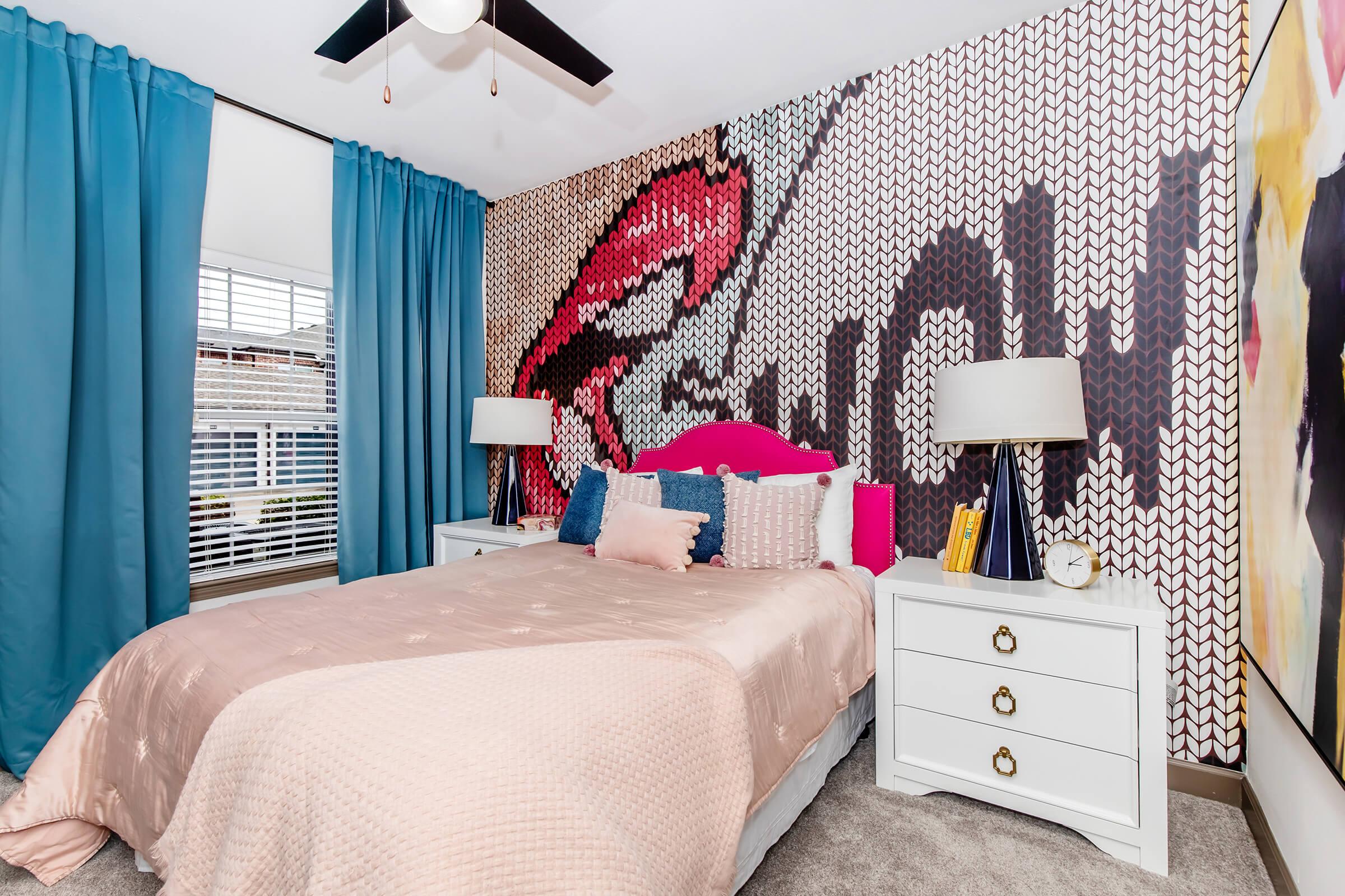 A stylish bedroom featuring a pink bedspread and vibrant accent pillows. The wall behind the bed has a bold graphic mural with the word "WOW." Two white nightstands with lamps are positioned on either side of the bed, and large blue curtains frame the window, adding a pop of color to the decor.