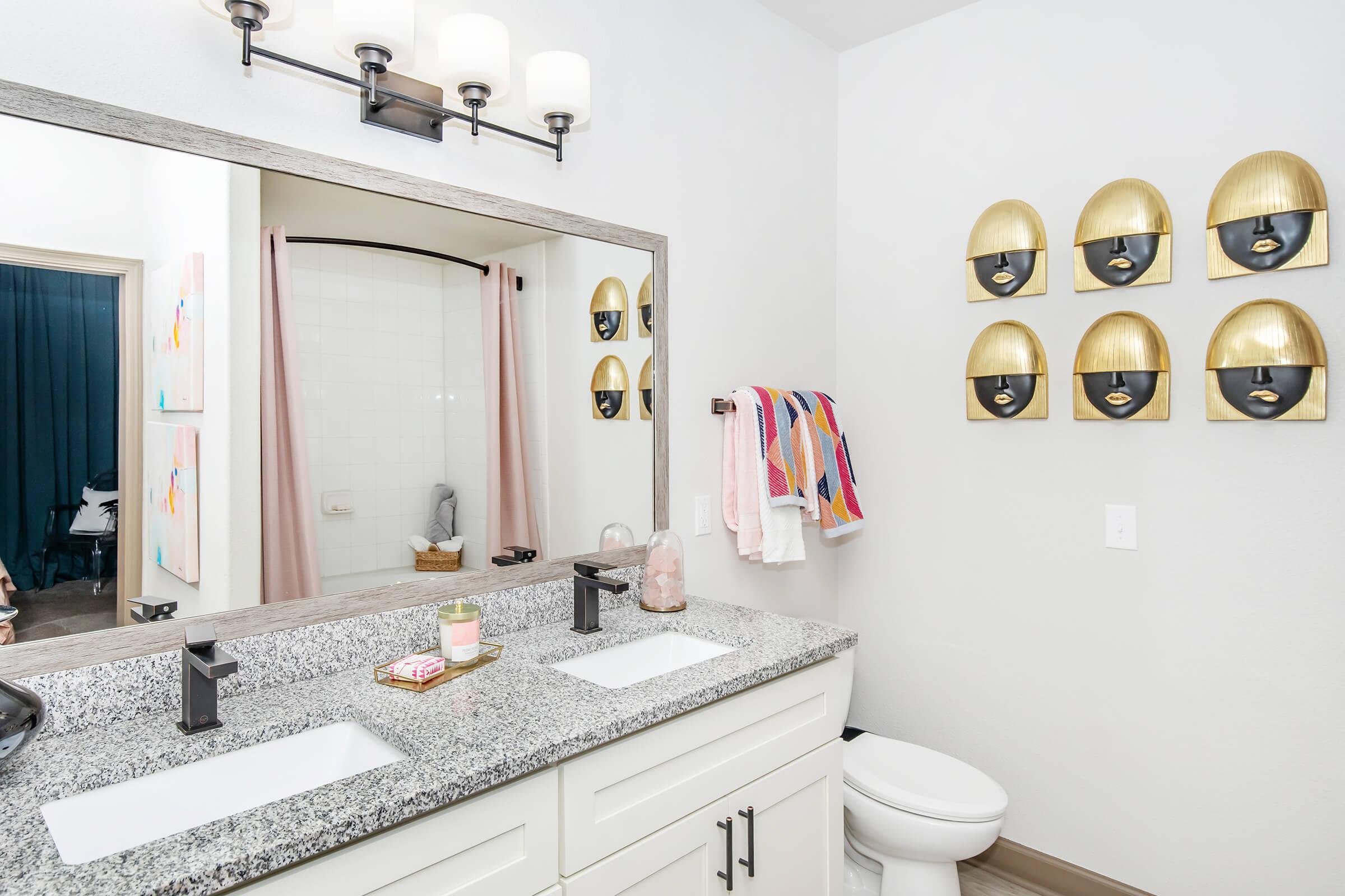 A modern bathroom featuring a double vanity with a granite countertop, black faucets, and a large mirror. The wall decor includes six gold wall sconces. A colorful towel is hanging on a rack, and a shower with a curtain is visible in the background, adding a touch of style to the space.