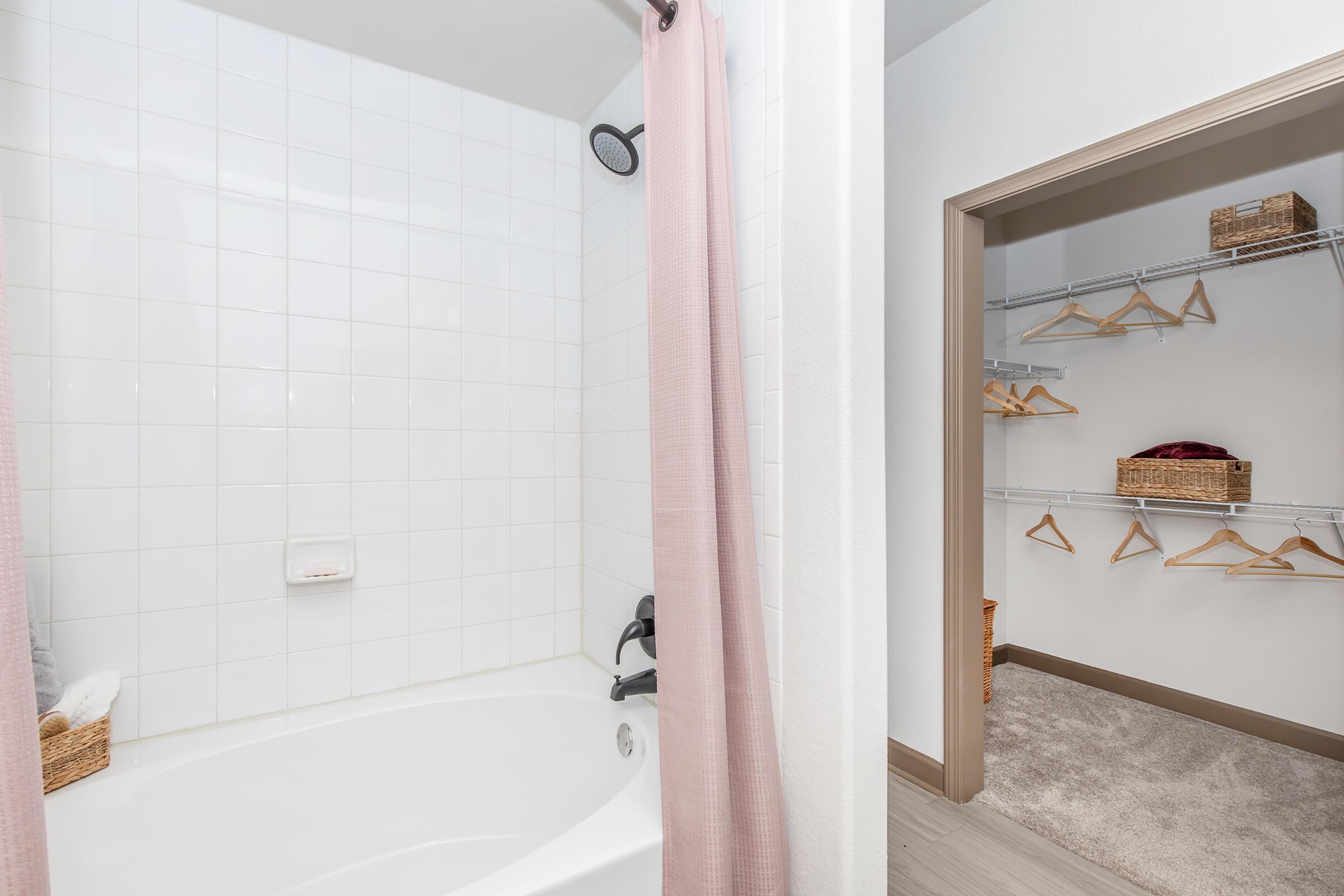 A clean bathroom featuring a shower with a pink curtain and a bathtub, along with a closet on the right. The closet has wooden shelves and hangers, with some baskets for storage. The overall aesthetic is neat and modern, with white tiles and light-colored walls.