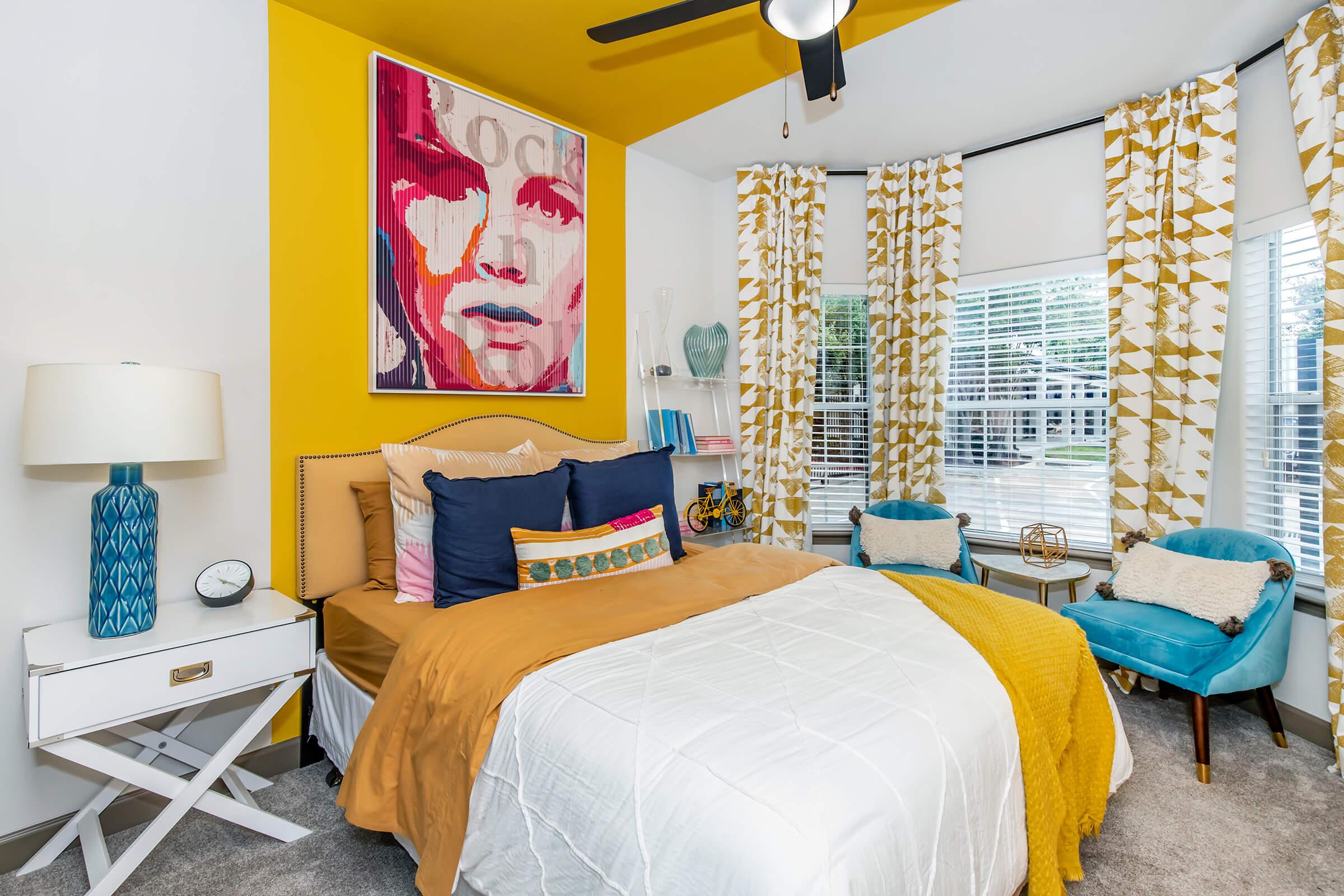 A cozy bedroom featuring a queen-sized bed with colorful pillows and a mustard-colored throw, a bright yellow accent wall with a pop art piece, and large windows draped with patterned curtains. A nightstand with a lamp and clock, along with two blue accent chairs, complete the inviting atmosphere.
