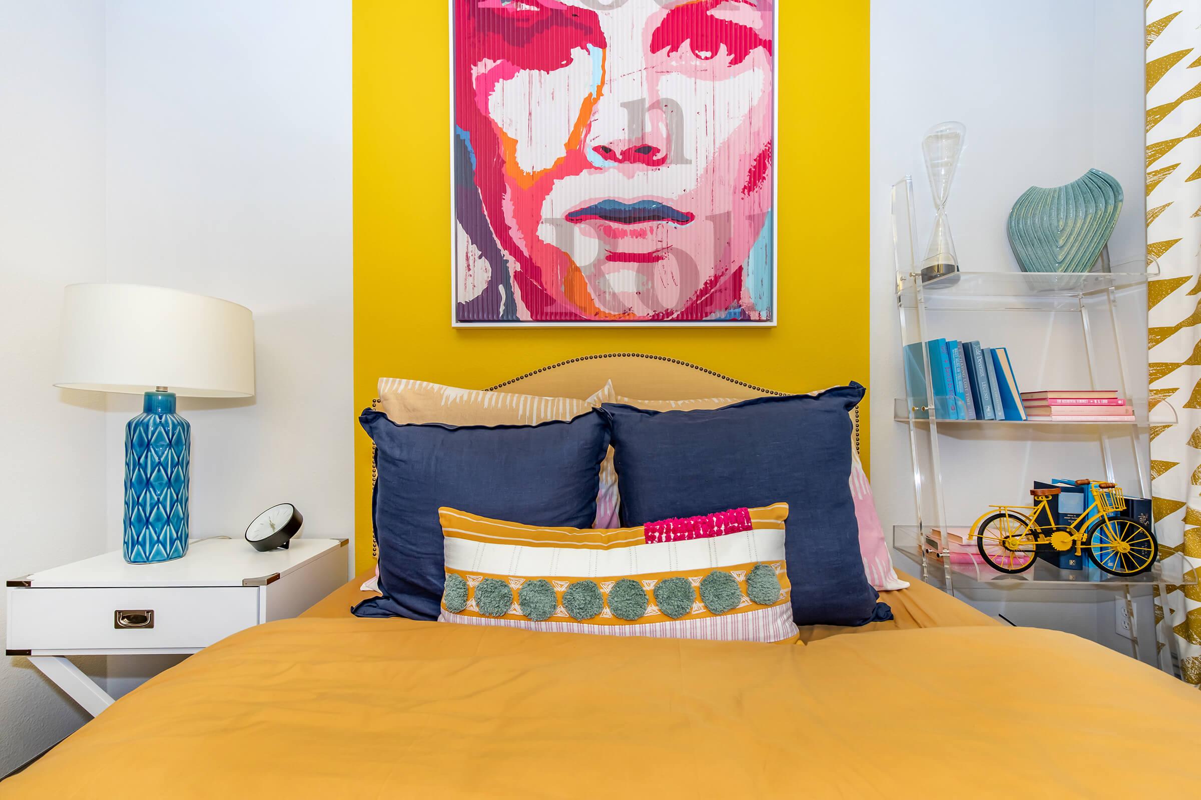 A cozy bedroom featuring a yellow wall with a vibrant portrait. The bed is dressed in a mustard-colored duvet with blue and patterned pillows. There's a white bedside table with a blue lamp and a clock. A clear shelving unit holds books and a decorative bicycle adds charm to the space.