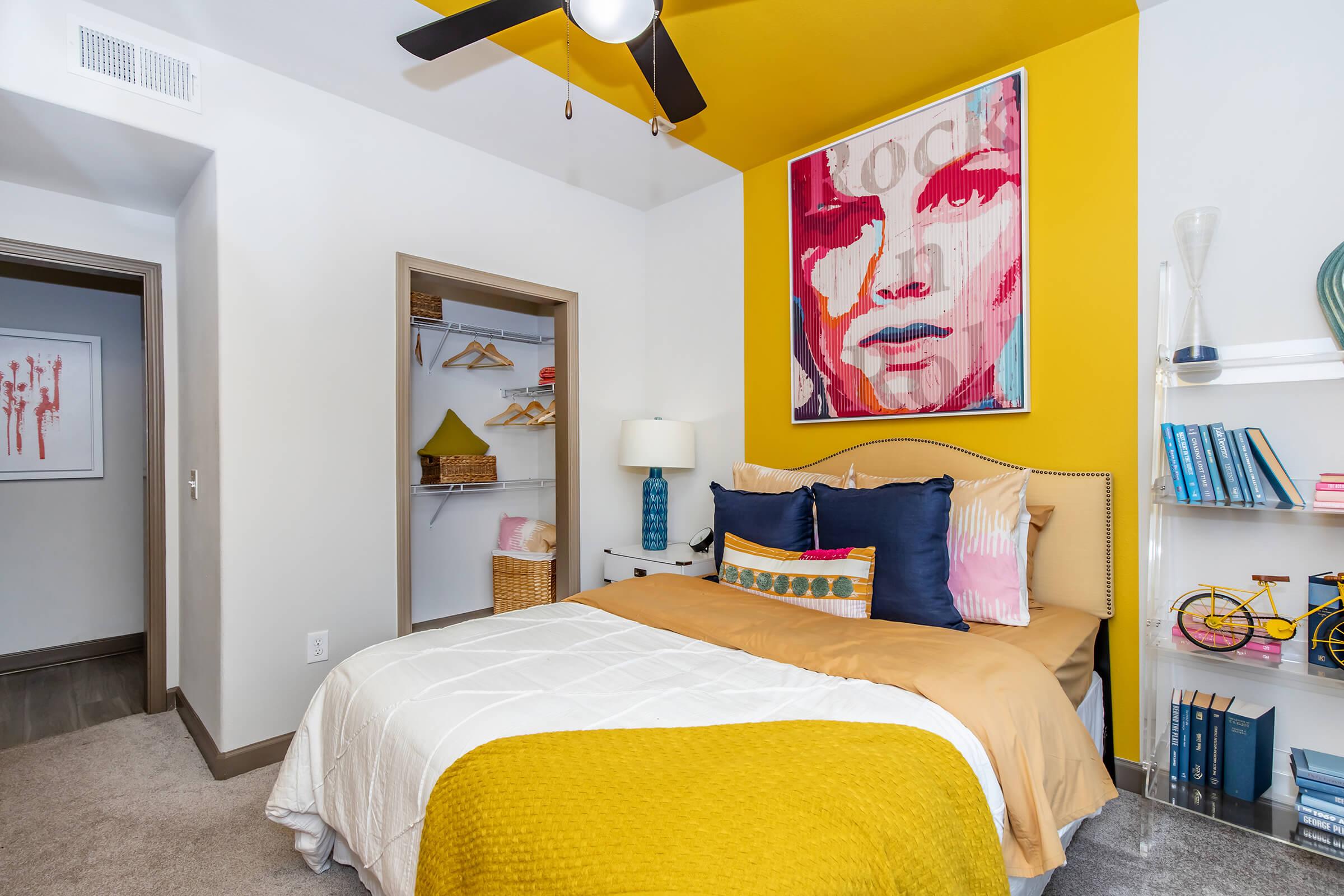 A cozy bedroom featuring a yellow accent wall with a bold artwork, a neatly made bed with colorful pillows and a textured blanket, a ceiling fan, and a small closet. Bookshelves and decorative items add a personal touch, while warm lighting creates an inviting atmosphere.