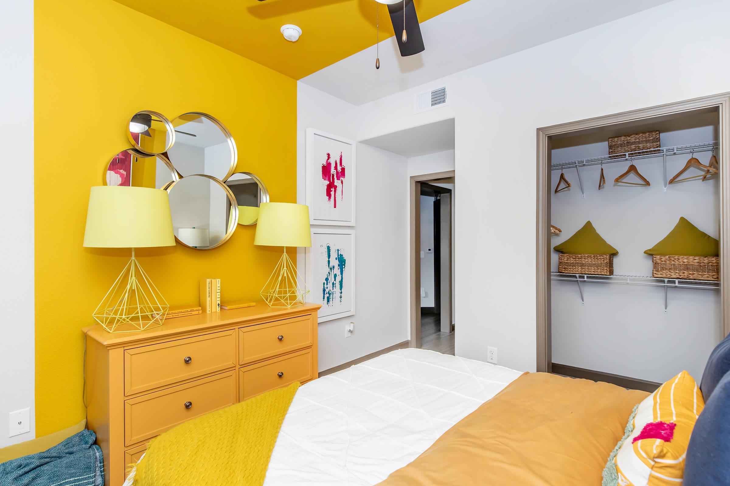 A cozy bedroom featuring a yellow accent wall and a ceiling fan. The room includes a stylish mustard dresser with geometric lamp stands, framed art on the wall, and a closet with hanging organizers. A white bedspread contrasts with mustard and green decorative pillows, creating a warm and inviting atmosphere.