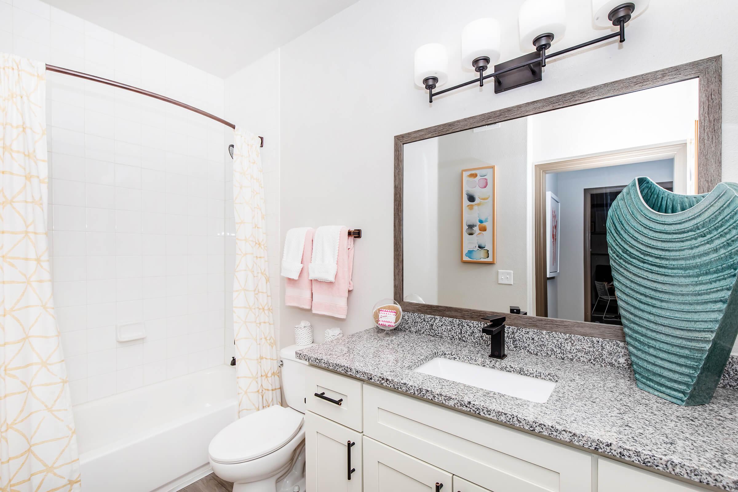 A modern bathroom featuring a white bathtub with a shower curtain, a granite countertop with a sink, a decorative vase, and a large mirror. Soft pink towels hang on the wall, and there is a framed artwork in the background, creating a clean and inviting atmosphere.