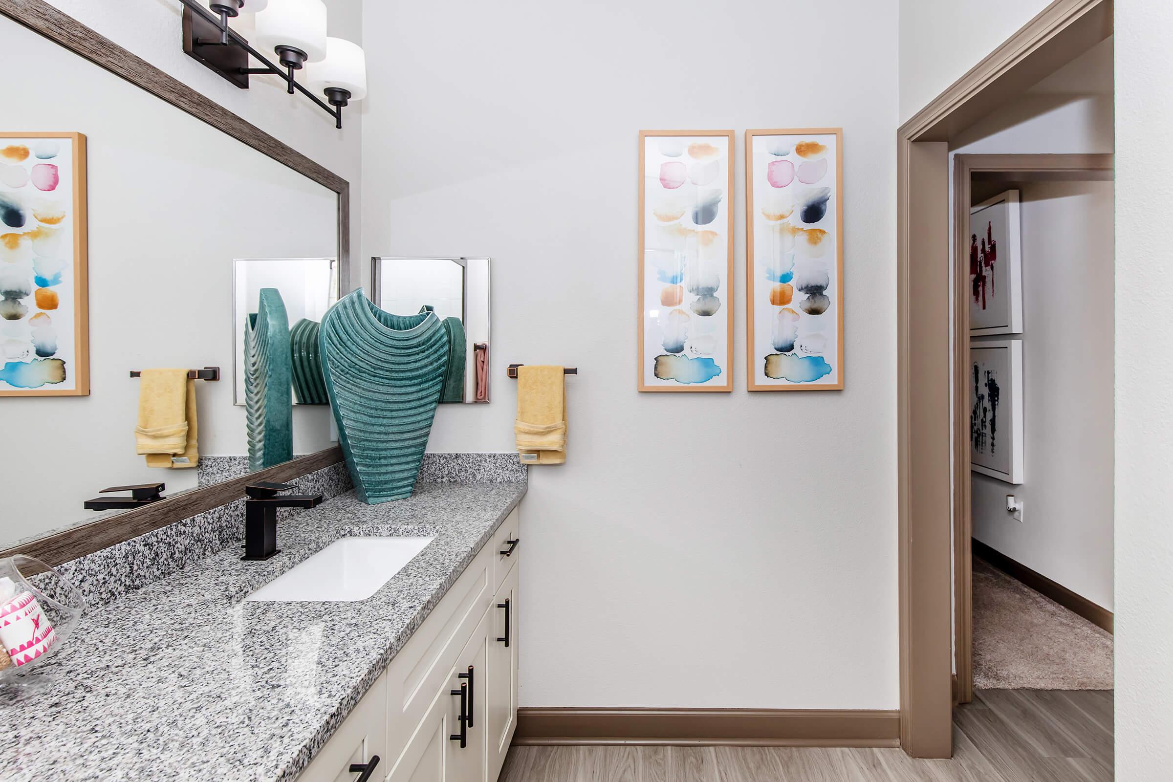 A modern bathroom featuring a granite countertop with a sink, a decorative turquoise vase on the left, and two colorful art pieces on the wall. Soft yellow towels hang beside the sink, and a mirror reflects the space, which is well-lit and has a warm, inviting ambiance. A doorway leads to another room.