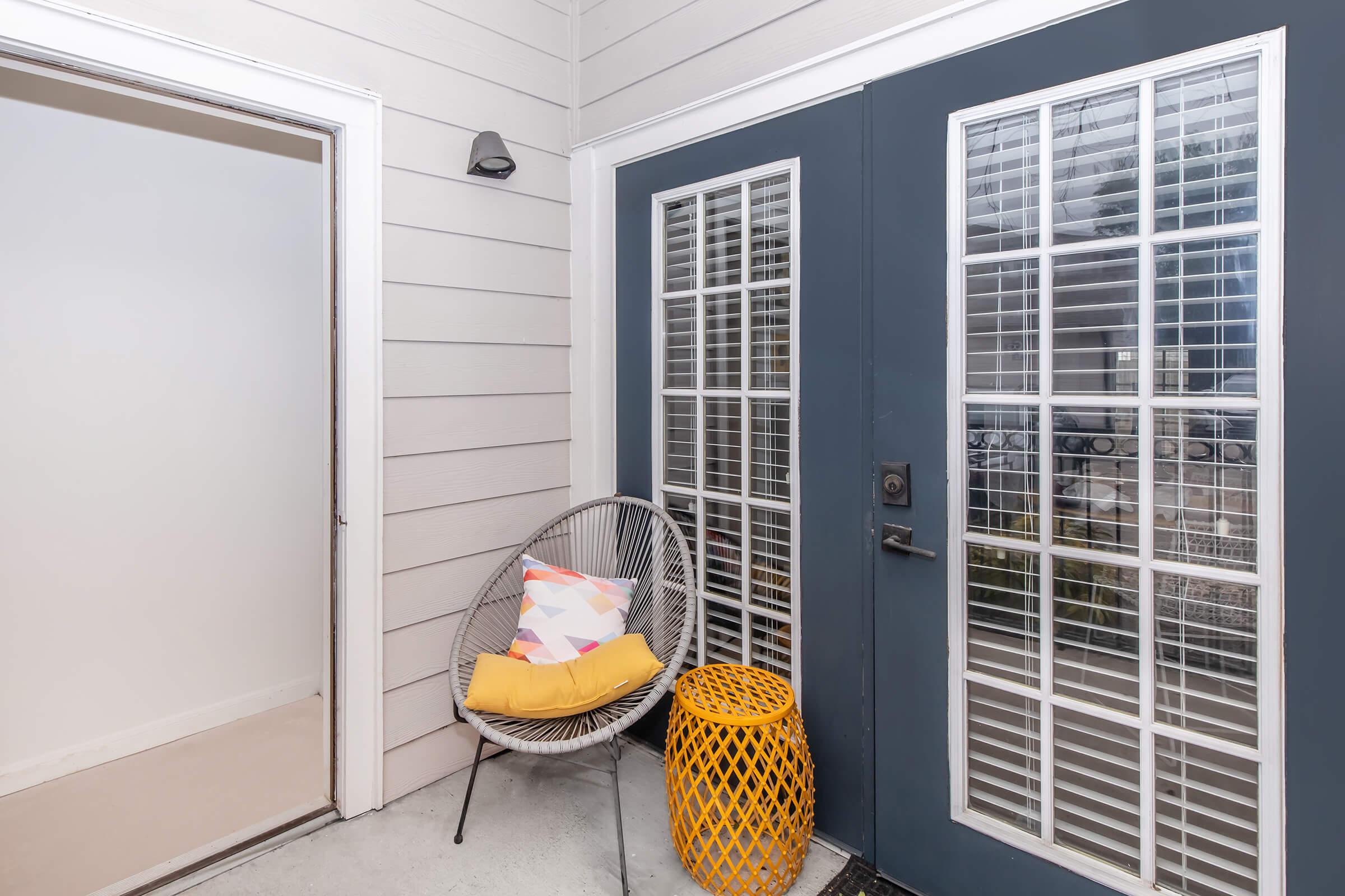 A cozy entryway featuring a pair of blue double doors with glass panes, a round yellow accent table, and a modern chair with a colorful cushion. The walls are light-colored wood, and the area has a clean, welcoming feel.