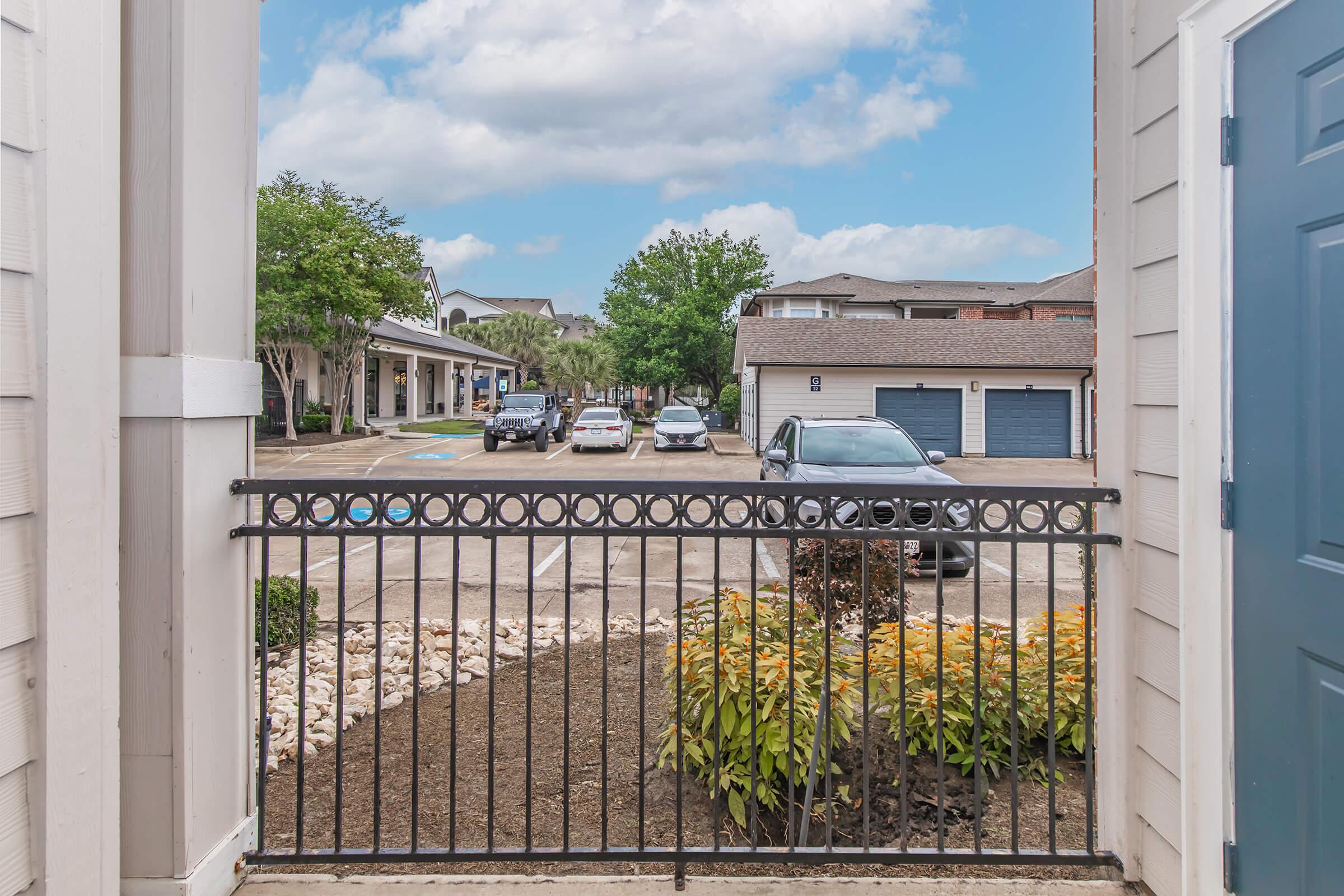 View from a gated entryway showing a parking lot with multiple vehicles. Surrounding greenery is visible, including shrubs and flower beds, with apartment buildings in the background under a cloudy sky.