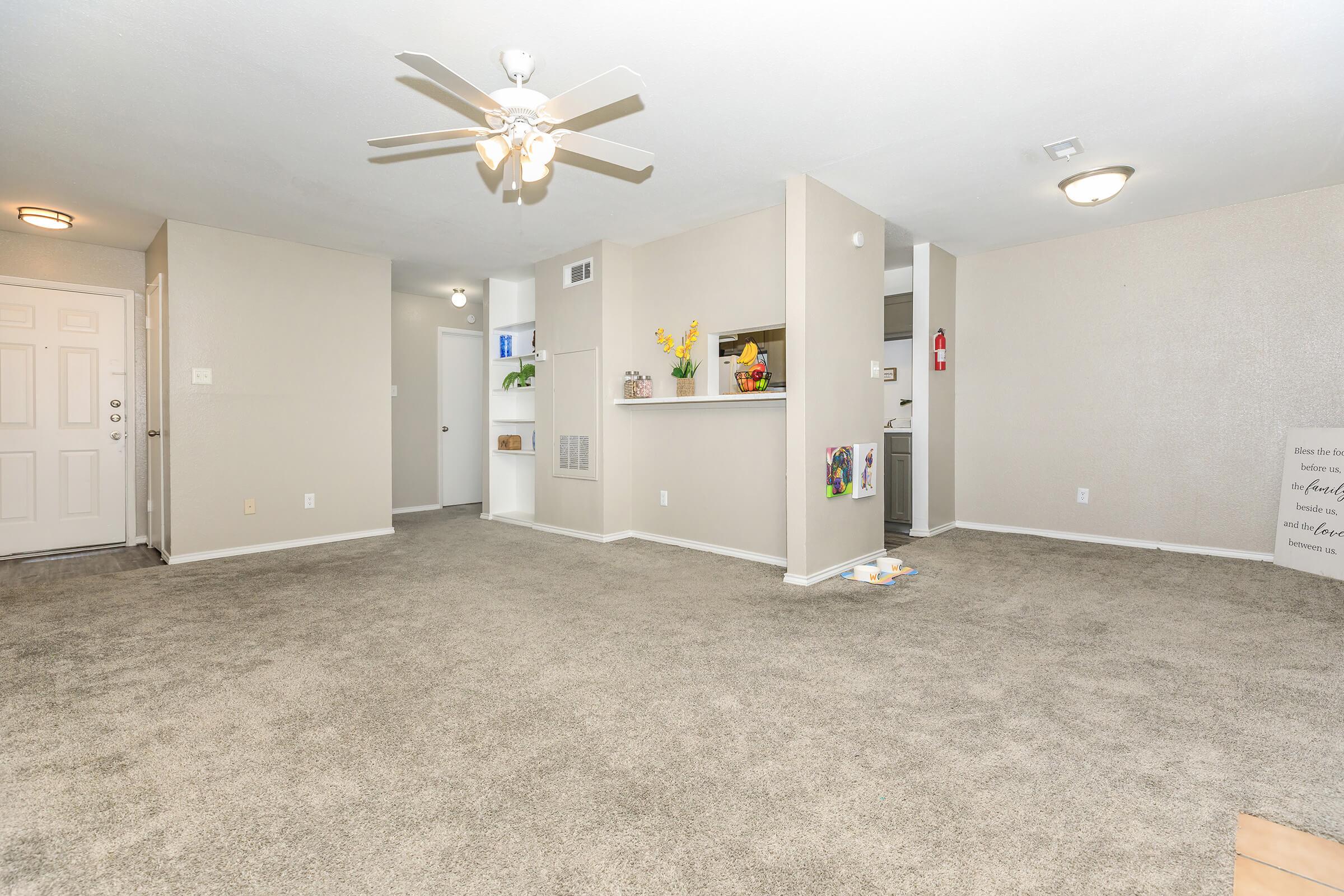 A spacious, well-lit living area with light grey carpet, featuring a ceiling fan. To the left, a front door and a small entryway. On the right, a partial wall with decorative items and shelves. The room is neutrally painted, creating a bright and inviting atmosphere, with no visible furniture.