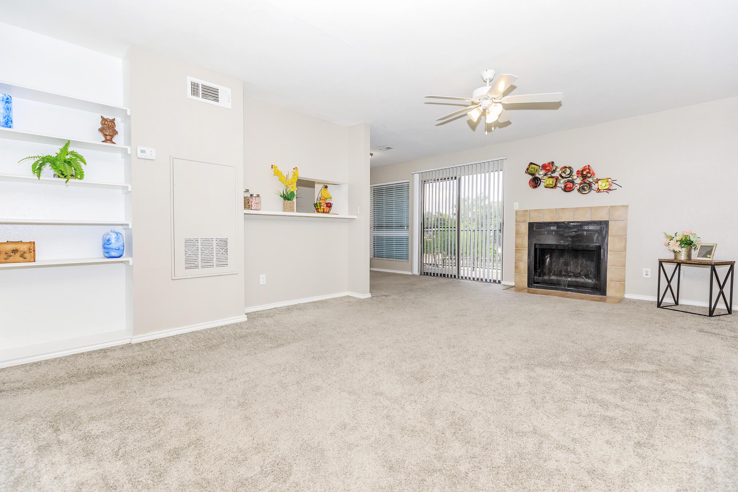 Spacious living room with beige carpet, featuring a ceiling fan, a fireplace, and a shelf with decorative items. Large windows with vertical blinds allow natural light, while a potted plant adds a touch of greenery. A side table is positioned next to the fireplace, enhancing the room's cozy atmosphere.