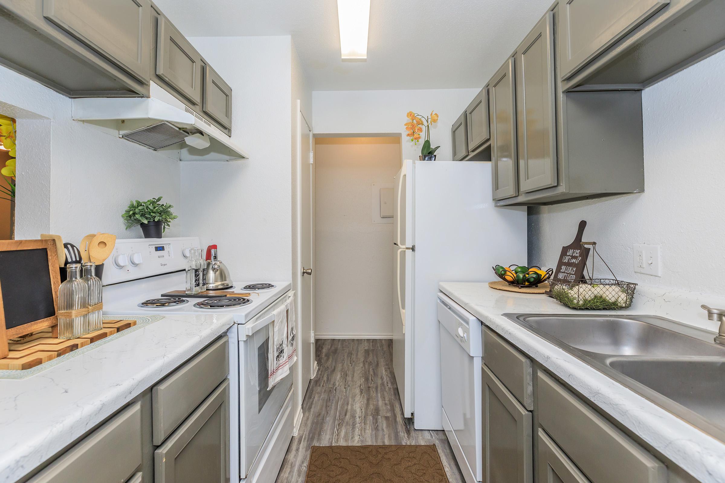 A modern kitchen featuring gray cabinets, a white stove, and a refrigerator. The countertop is made of light-colored stone, and there is a sink with a dish rack. Decorative items like a kettle and a bowl of fruit are present, along with potted plants and a small welcome sign. The space is bright and cozy.