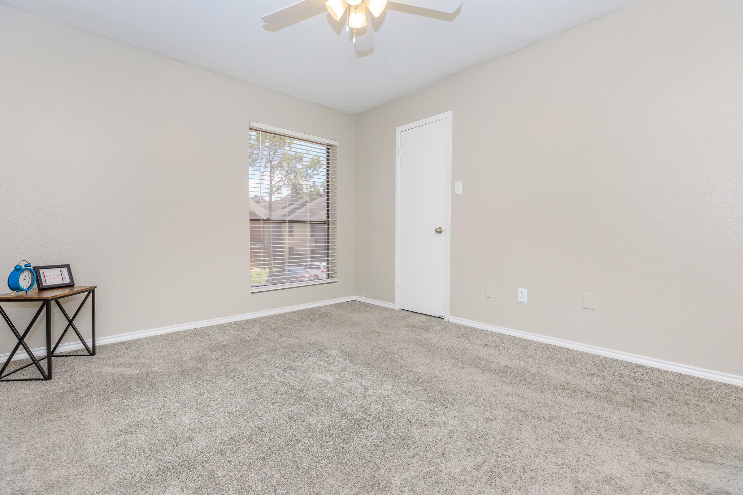 A bright, empty room with light gray walls and a plush carpet. There's a window with blinds letting in natural light, a ceiling fan, and a closed white door. A small table with a decorative blue clock and a framed picture is in one corner, enhancing the simple, clean aesthetic.