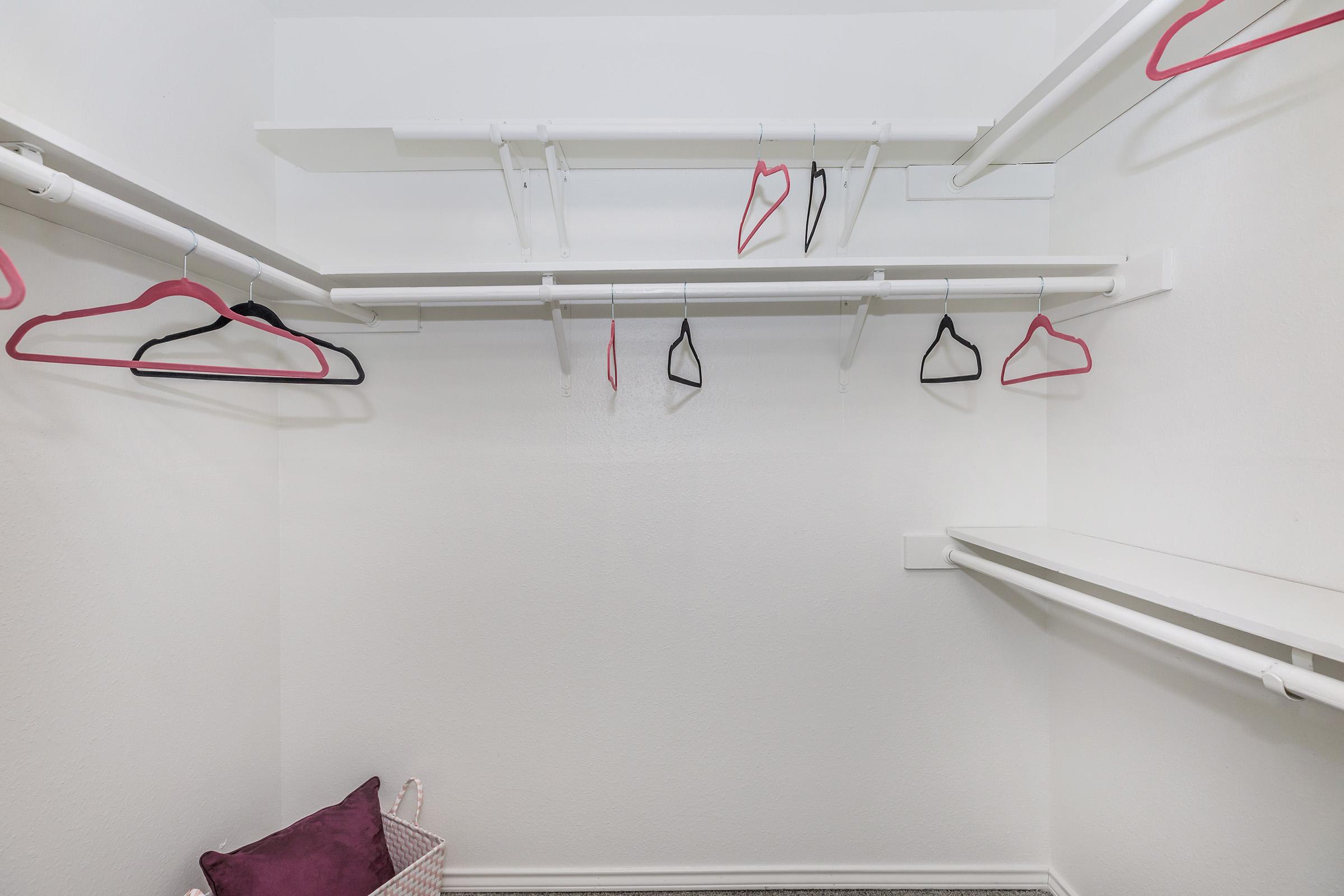 An empty closet with several hanging clothes hangers on white rods. A small basket is placed in the corner. The walls are plain and white, and the floor is carpeted in a neutral color.
