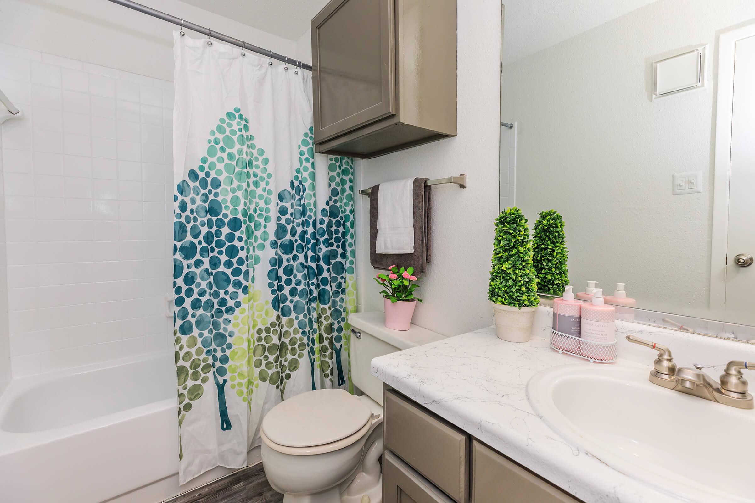 A modern bathroom featuring a white bathtub with a colorful tree-patterned shower curtain, a stylish countertop with potted plants and toiletries, and gray cabinetry above. The space is well-lit, with a mirror and a towel rack visible, creating a fresh and inviting atmosphere.