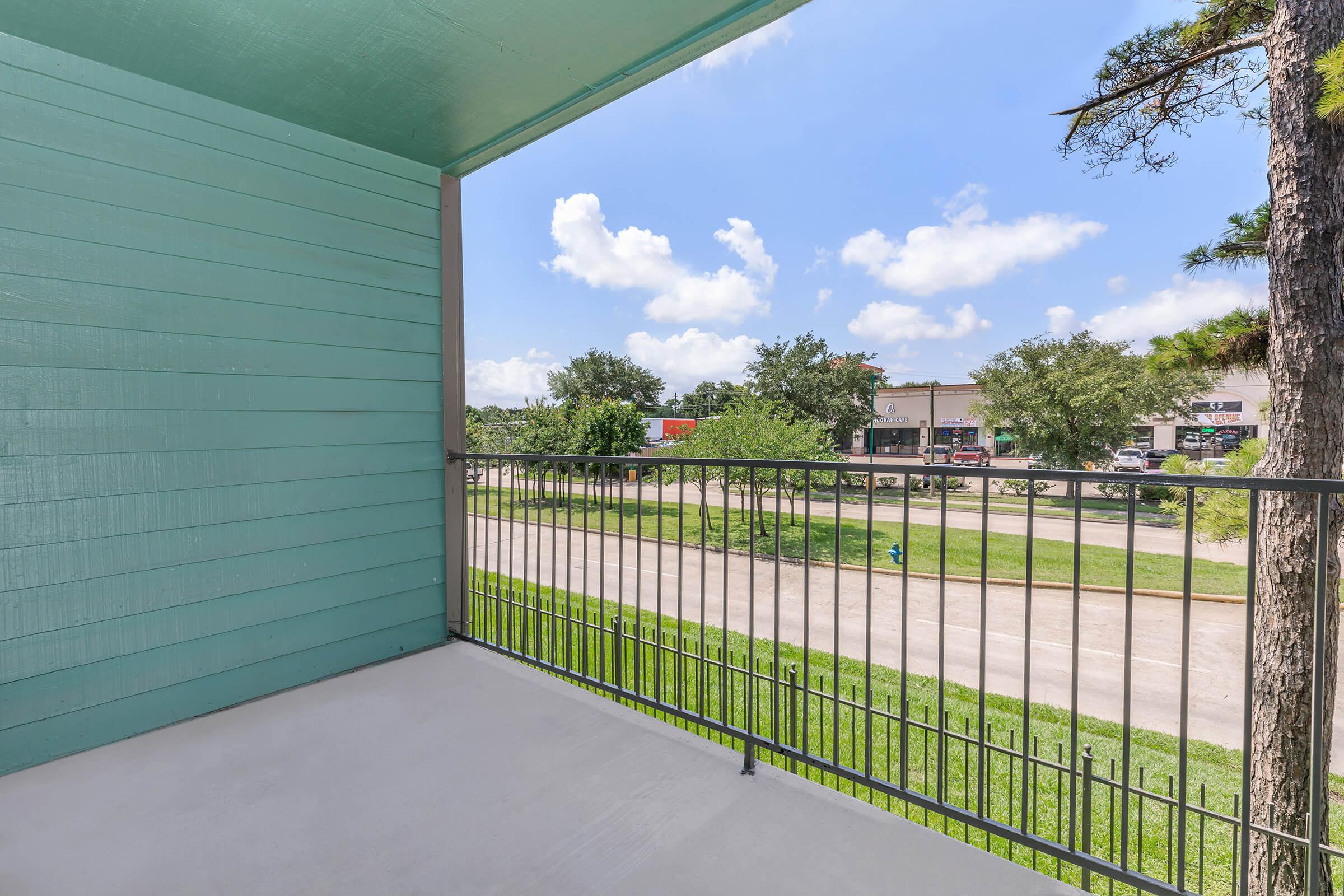 A view from a balcony featuring a green-painted wall and railings. In the background, there are trees and buildings along a street under a partly cloudy sky. The scene conveys a bright, open outdoor space.
