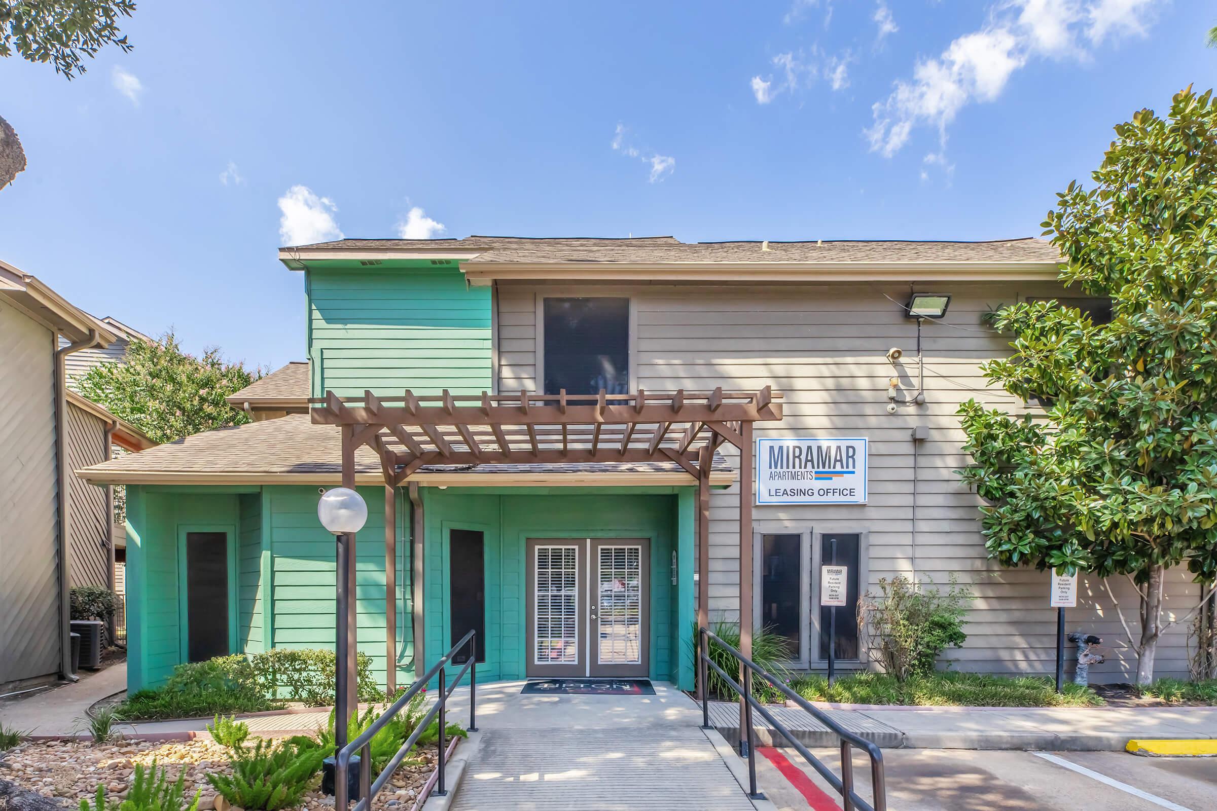 A leasing office building with a wooden pergola at the entrance. The exterior features a combination of green and beige siding. The office has large windows and a sign displaying "MIRAMAR Leasing Office." Surrounding greenery and a paved walkway add to the welcoming appearance. A clear blue sky can be seen above.