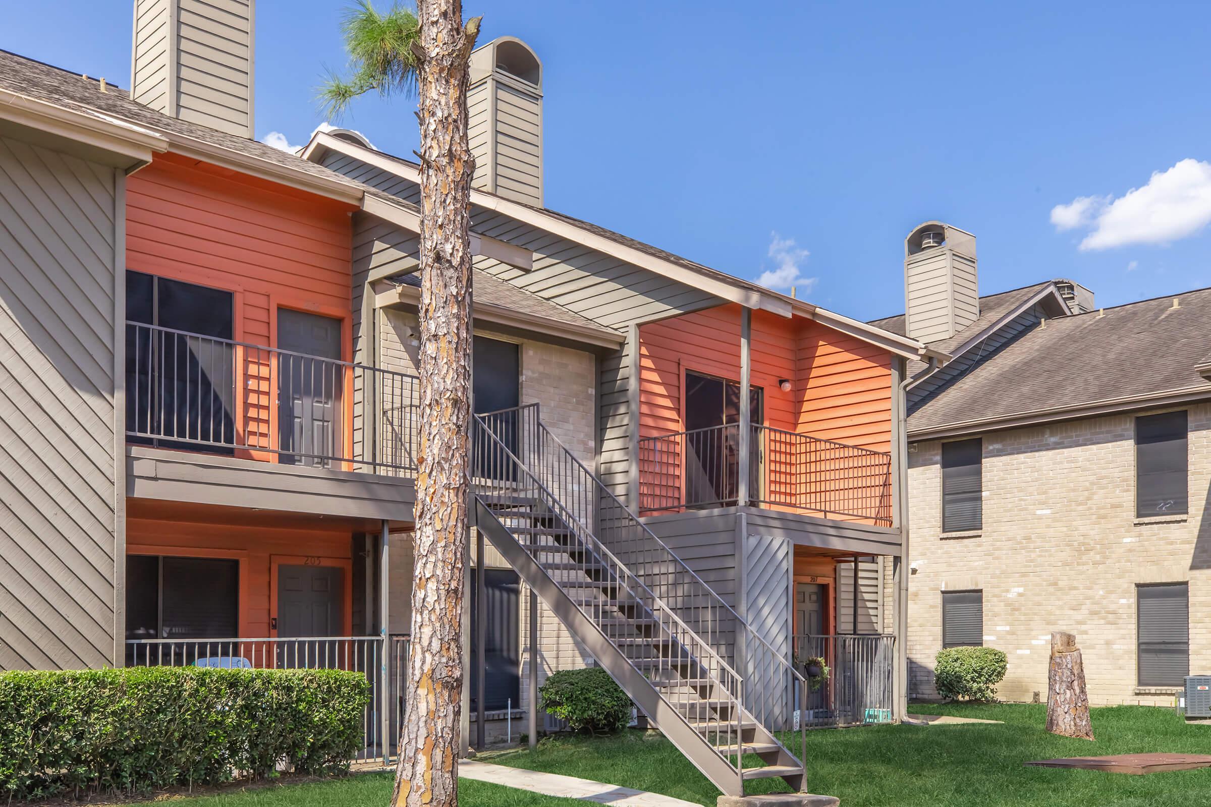 A multi-story apartment complex featuring a staircase leading to an upper balcony. The building has a combination of orange and gray siding, with greenery and blue skies in the background. The scene captures a sunny day, highlighting the architectural design and landscape of the area.