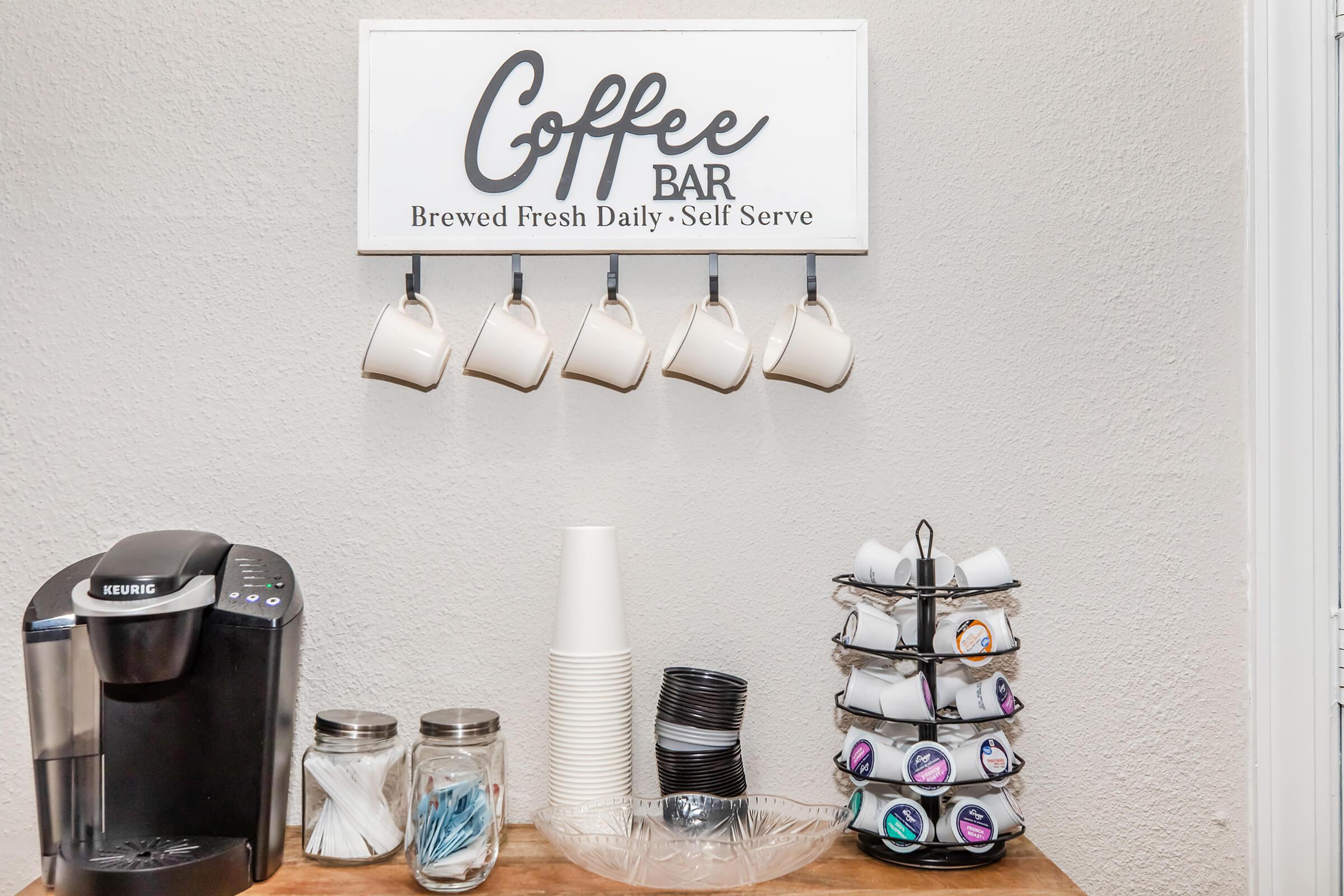 A coffee bar setup featuring a sign that says "Coffee Bar" with the subtitle "Brewed Fresh Daily - Self Serve." There's a Keurig coffee maker, several stacked cups, jars with straws and stirrers, and a carousel displaying various coffee pods.