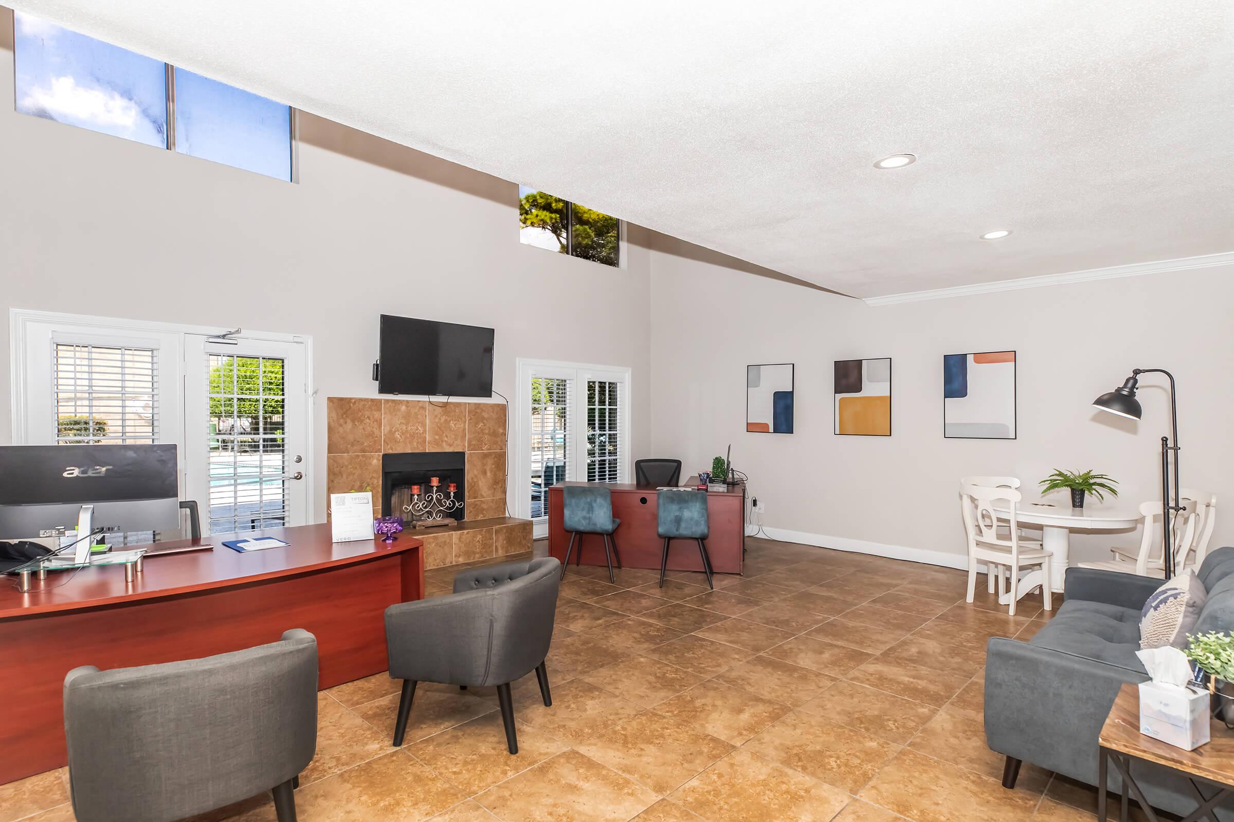 Interior of a modern reception area featuring a front desk, chairs, and a cozy seating arrangement. A fireplace is visible on one wall, with a television mounted above it. Large windows bring in natural light, and artwork decorates the walls. The space is designed for comfort and functionality.