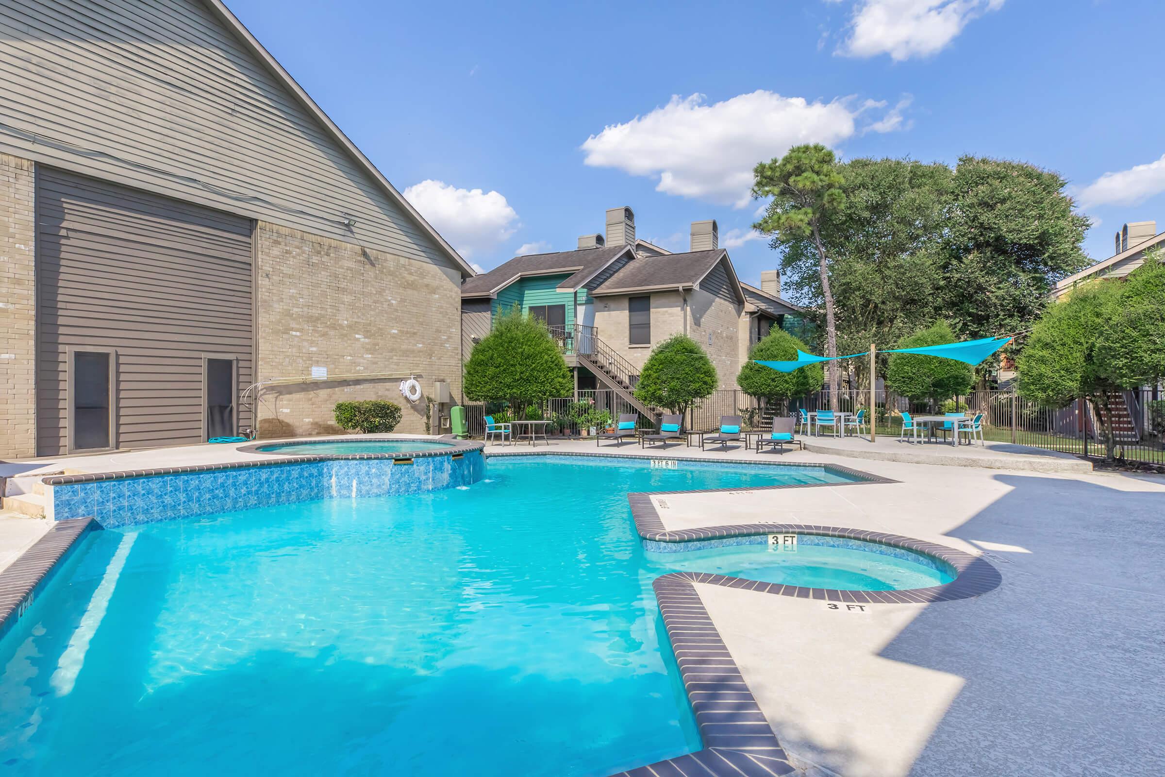 A bright outdoor swimming pool surrounded by lounge chairs and neatly trimmed bushes. There are two hot tubs adjacent to the pool, with sunny blue skies and a few clouds overhead. In the background, apartment buildings are visible, creating a relaxing residential atmosphere.