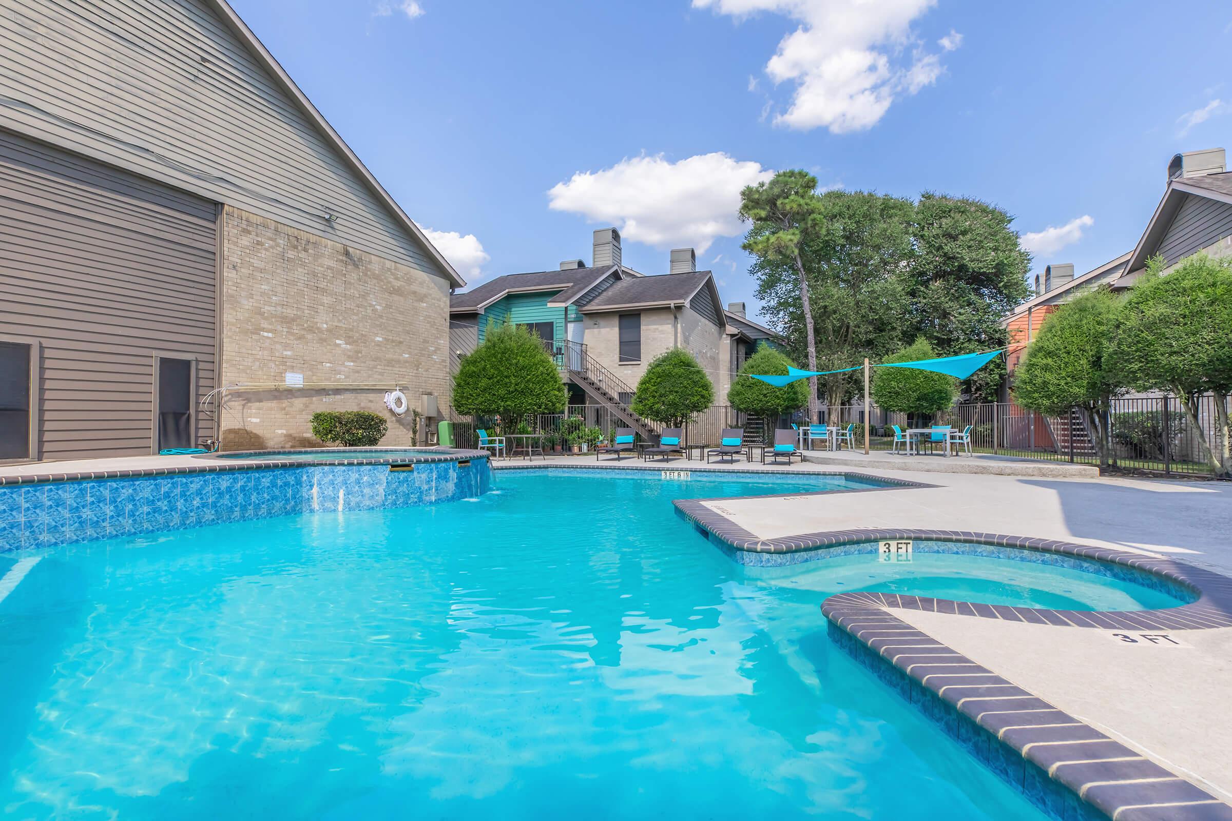 A swimming pool surrounded by apartment buildings. The pool features a spa area and is bordered by lounge chairs and umbrellas. Lush green trees and shrubs provide some shade, while a clear blue sky with scattered clouds adds to the serene atmosphere.