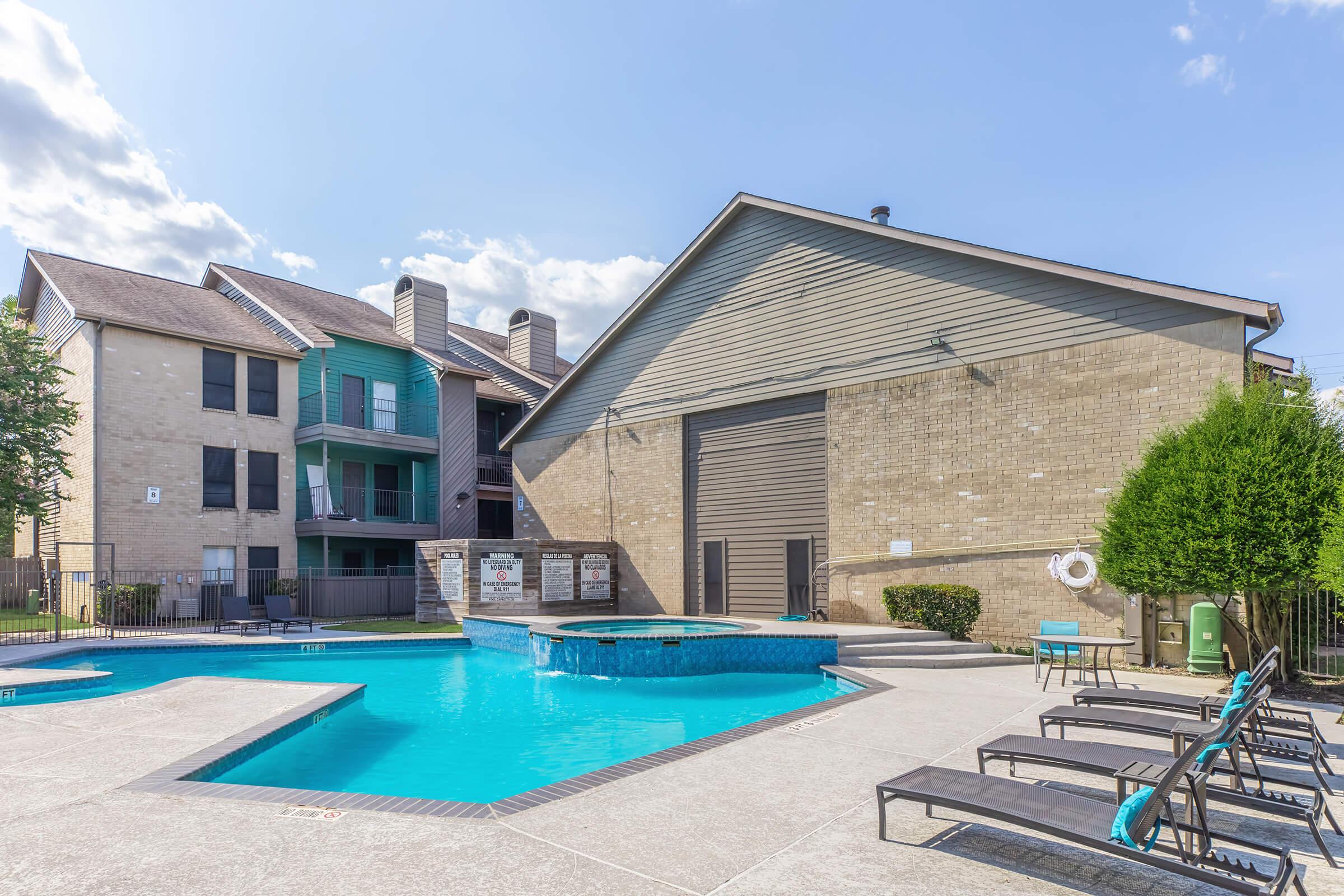 A sunny view of an outdoor swimming pool area with a clear blue pool, surrounded by lounge chairs. In the background, there are residential buildings with a mix of brick and green siding, and well-manicured landscaping. The sky is bright with a few clouds.