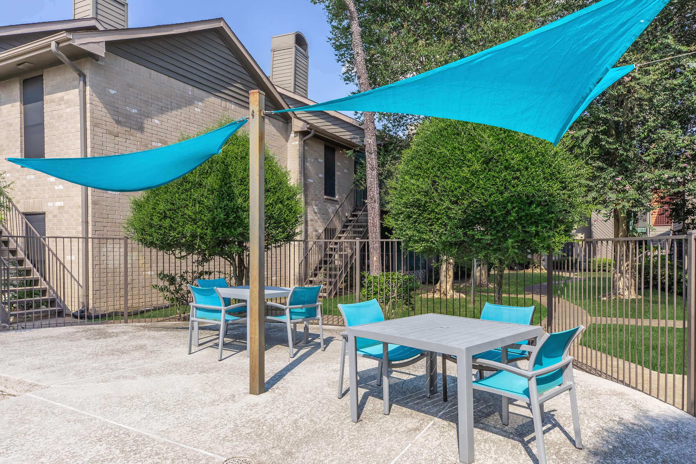 A patio area featuring light blue shade sails overhead, surrounded by greenery. The space includes a gray table and several turquoise chairs, set on a concrete surface. In the background, there are stairs leading to two buildings. It's a sunny day with clear blue skies.