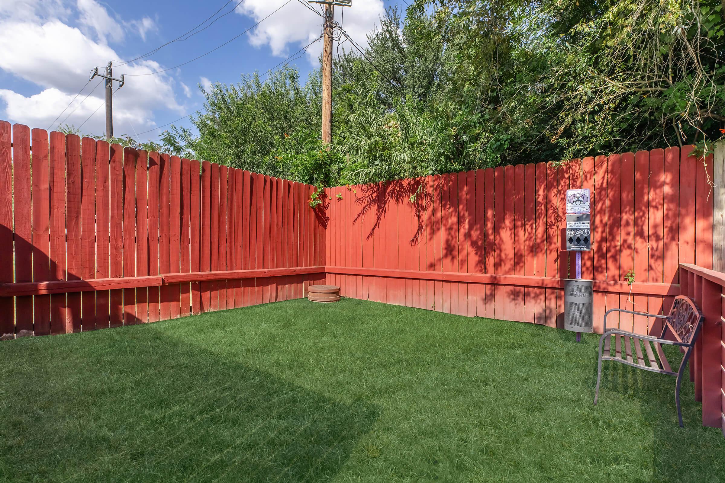 A small outdoor space with a neatly trimmed grassy area surrounded by tall red wooden fences. One corner features a metal bench, while a small cylindrical table is visible nearby. The scene is bright, with blue skies and fluffy clouds above, creating a pleasant and secluded atmosphere.