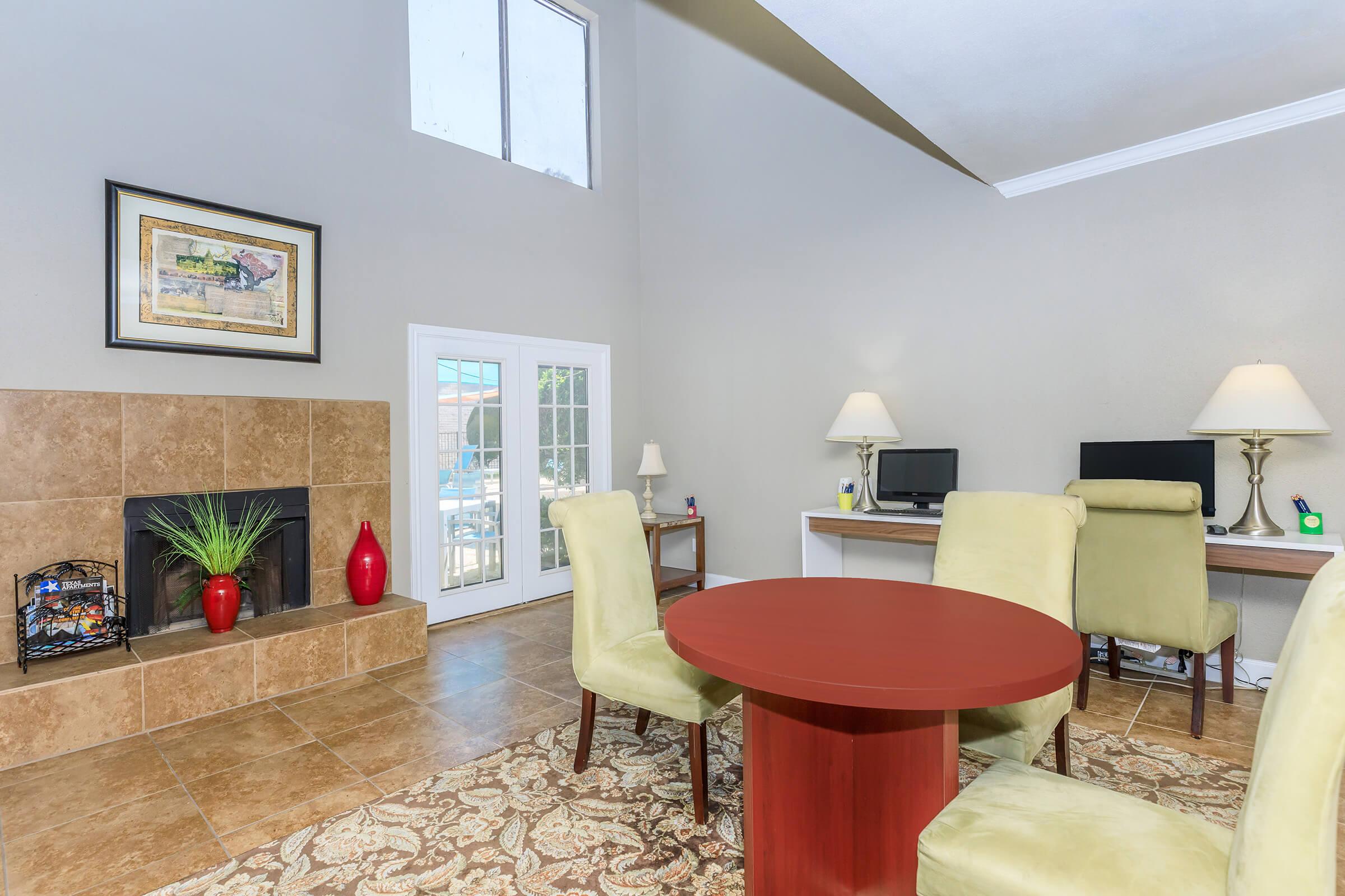 A spacious living room featuring a cozy fireplace, two computer desks with lamps, and a round red dining table surrounded by green chairs. Large windows allow natural light to fill the room, and the decor includes a framed painting and decorative vases. A soft area rug complements the tiled floor.