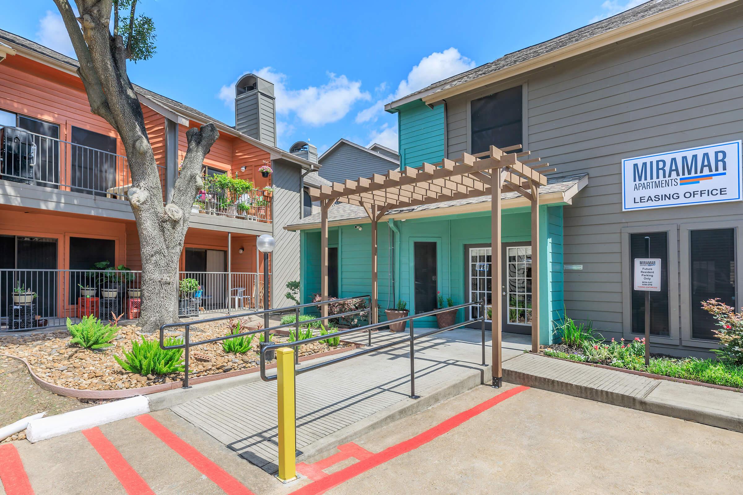 Leasing office entrance of Miramar Apartments featuring a teal building with a sign, a pergola, and accessible ramp. Surrounding landscaping includes rocks, plants, and small trees, with other apartment buildings visible in the background under a partly cloudy blue sky.
