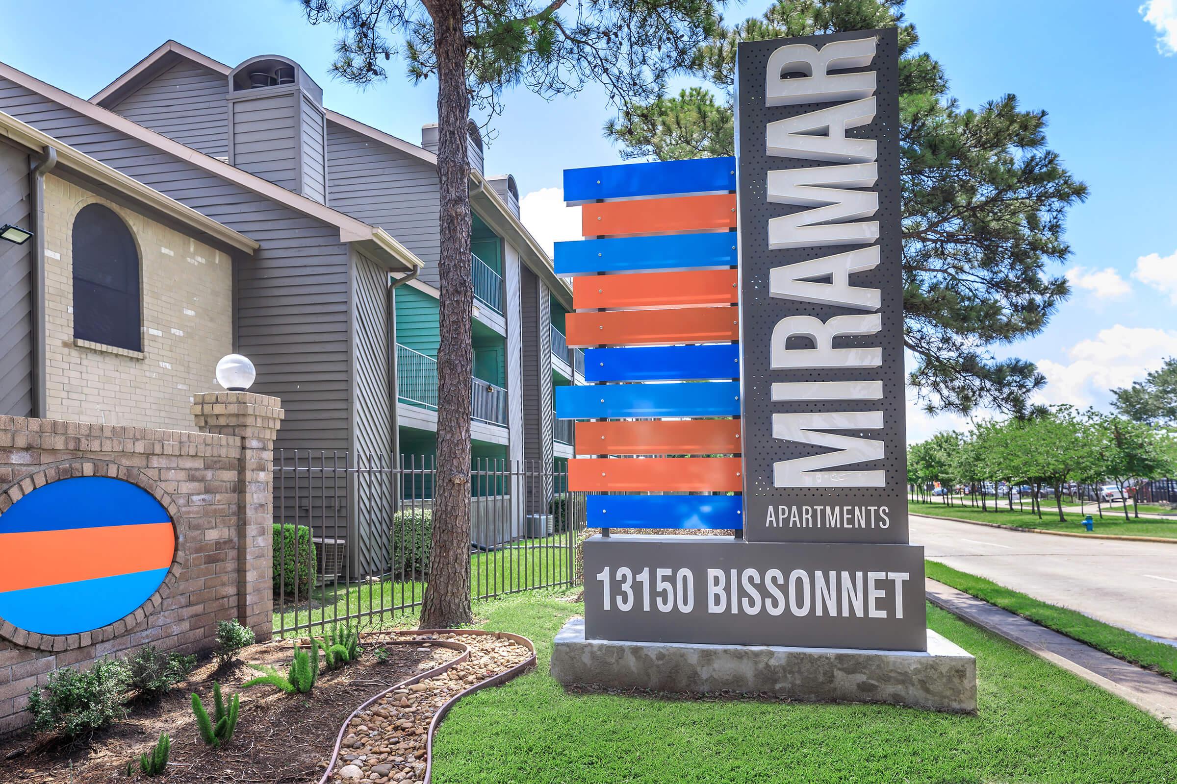 Sign for Miramar Apartments located at 13150 Bissonnet. The sign features a modern design with blue and orange horizontal stripes and the apartment name prominently displayed. Surrounding the sign are green lawn areas, including small shrubs and trees.