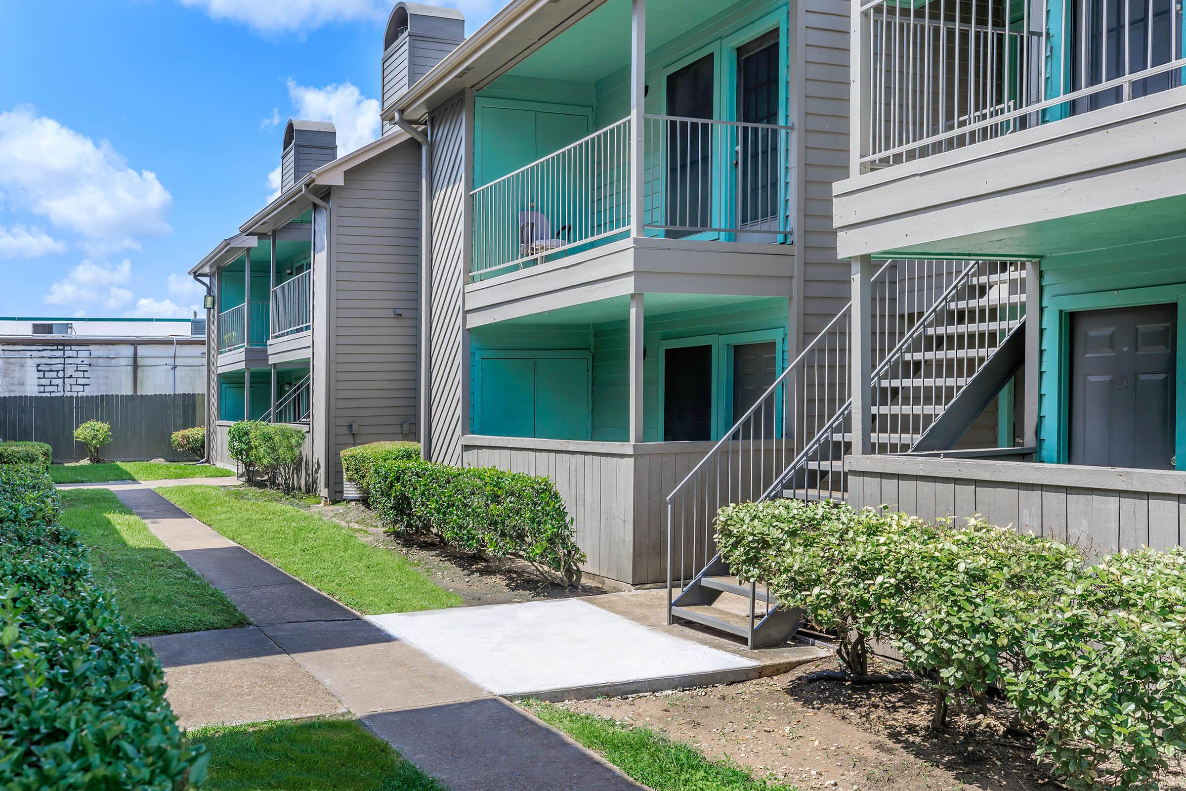 A view of a two-story apartment complex featuring green balconies, a connecting staircase, and well-maintained landscaping with shrubs and grass. The walkway leads through the courtyard with a clear blue sky above.