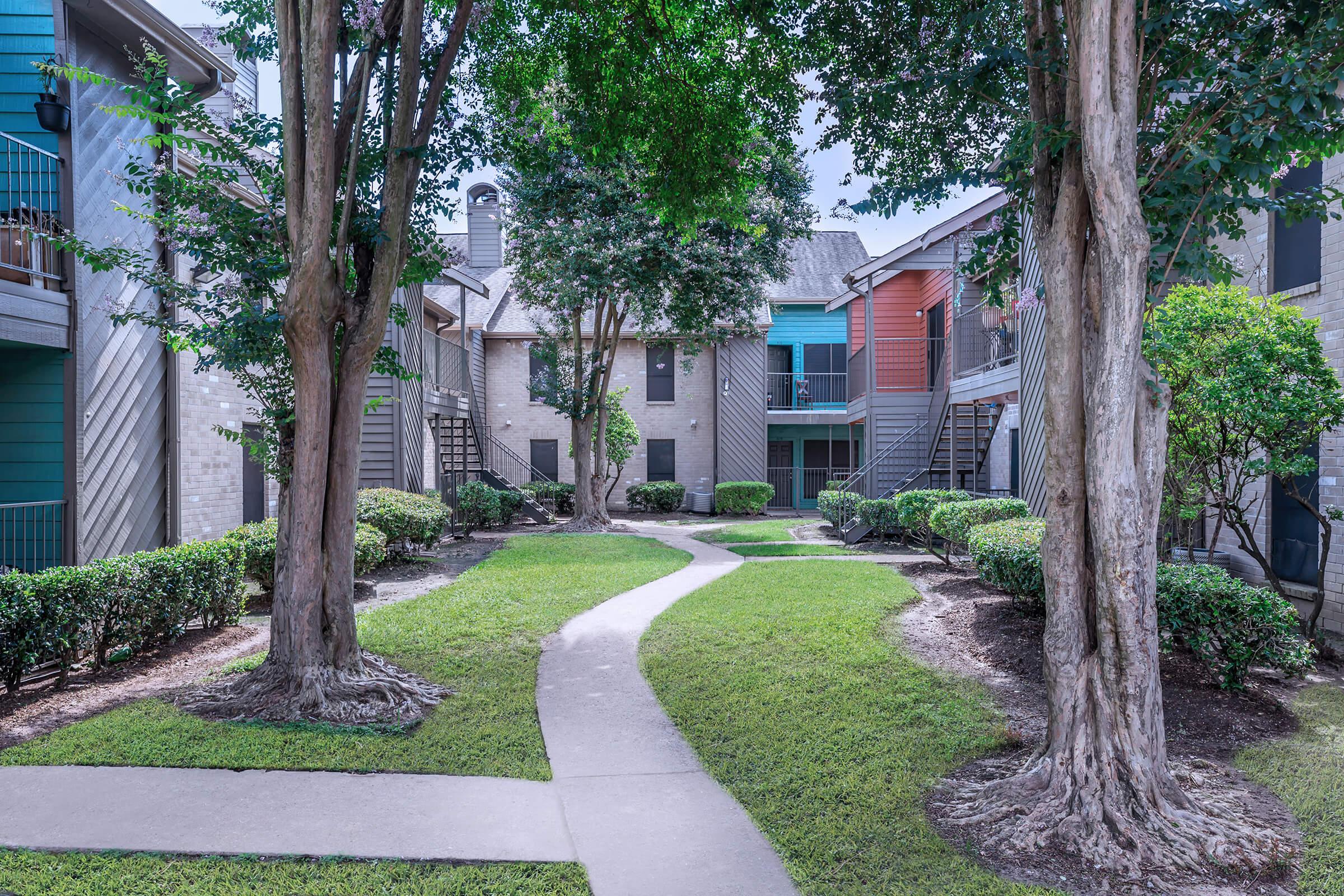 A view of a landscaped courtyard featuring a winding path that leads through grassy areas and shrubs. Surrounding the path are multi-story apartment buildings in varying colors, with trees providing shade. The scene is serene and well-maintained, inviting for residents and visitors alike.