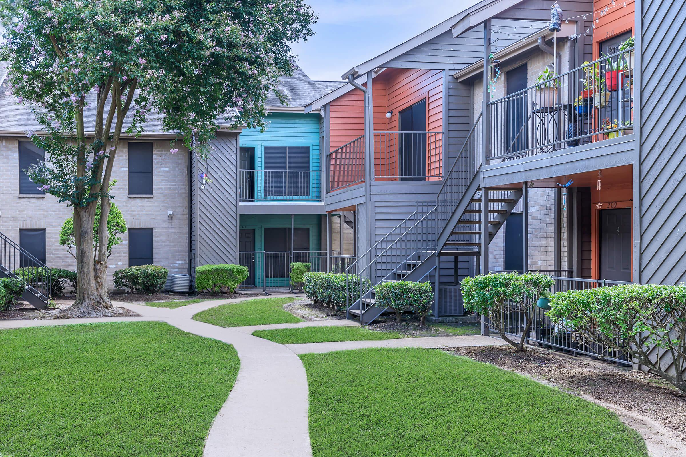 A view of a residential apartment complex featuring two buildings with colorful exteriors, a well-maintained lawn, and paved walkways. The buildings have balconies and stairs visible, with greenery surrounding the area, creating a serene atmosphere.