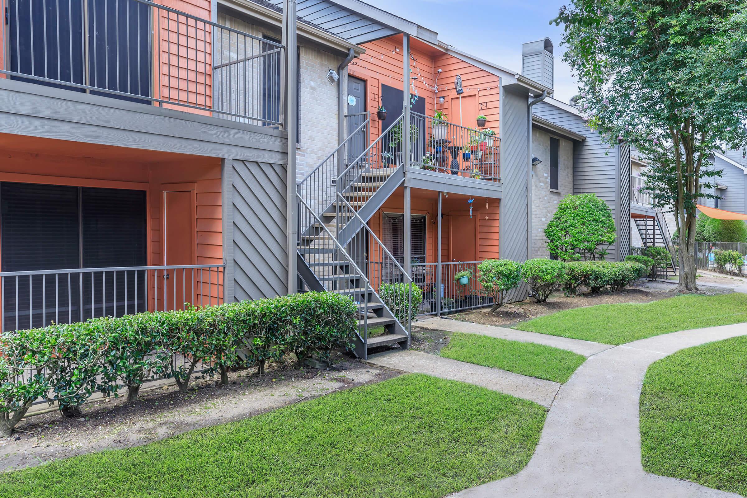 Exterior view of a residential apartment complex featuring multiple units, some with orange accents. There are balconies, a staircase leading to upper floors, and well-maintained greenery with manicured hedges and lawn around a curving pathway. Bright, clear sky above adds to the inviting atmosphere.