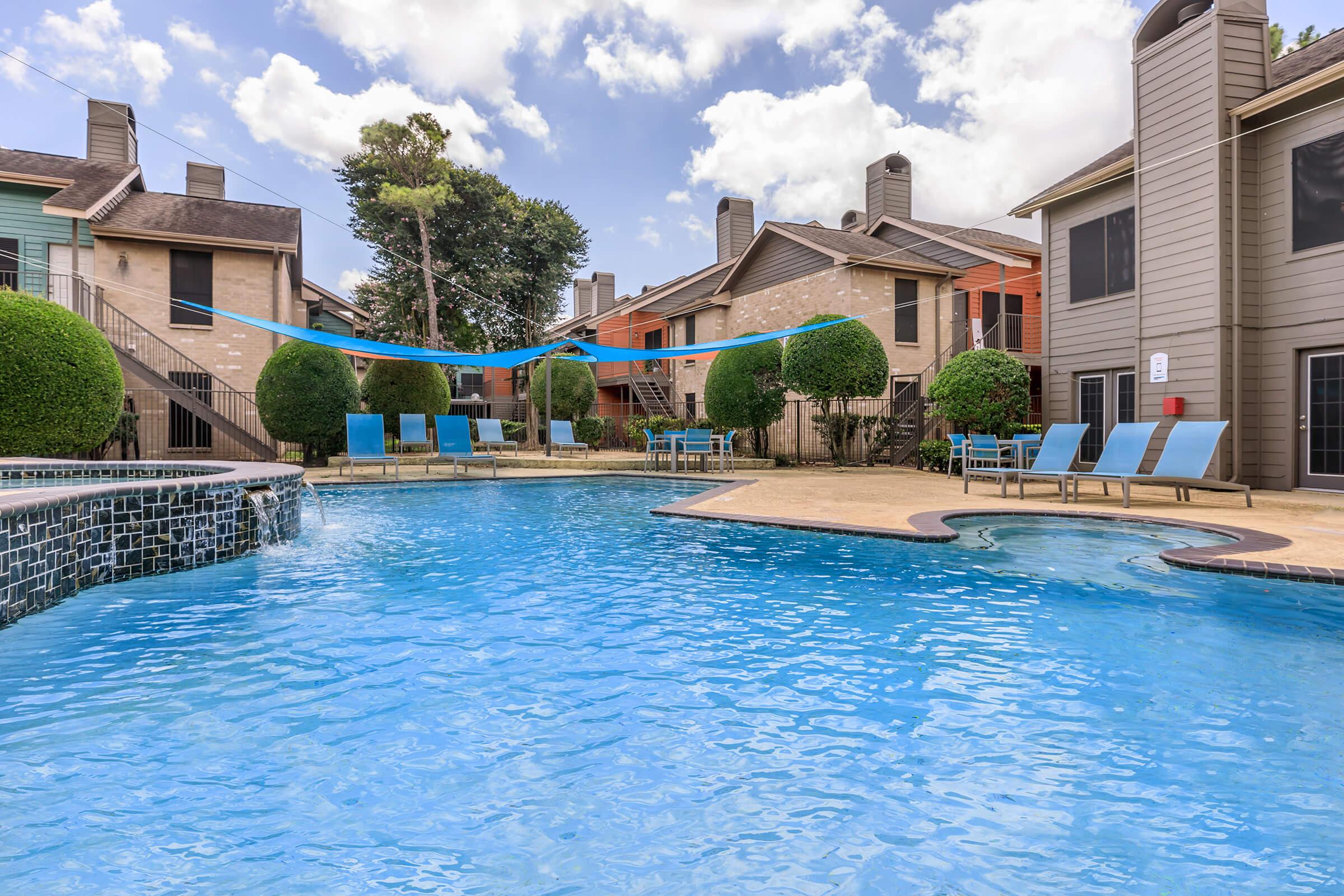 A swimming pool surrounded by lounge chairs and neatly trimmed hedges, with colorful buildings in the background under a partly cloudy sky. A blue ribbon decoration spans across the pool area, creating a vibrant atmosphere.