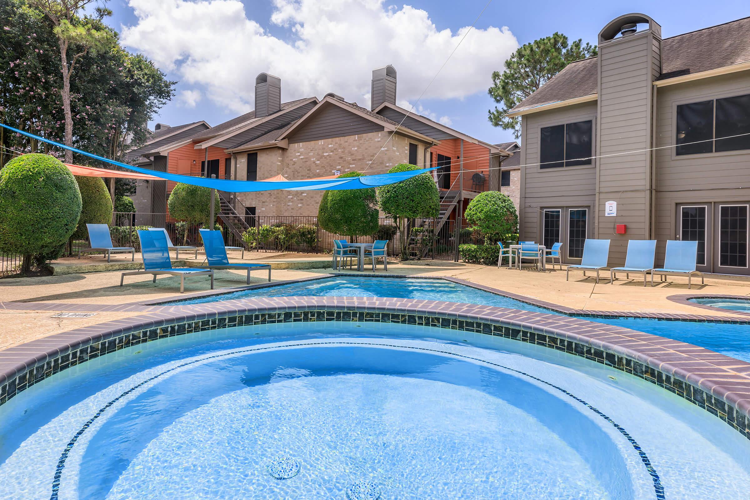 A sunny pool area with a sparkling blue pool and spa in the foreground. Comfortable lounge chairs surround the pool, and colorful shade sails provide protection from the sun. In the background, well-maintained apartment buildings are visible, surrounded by greenery and trees.