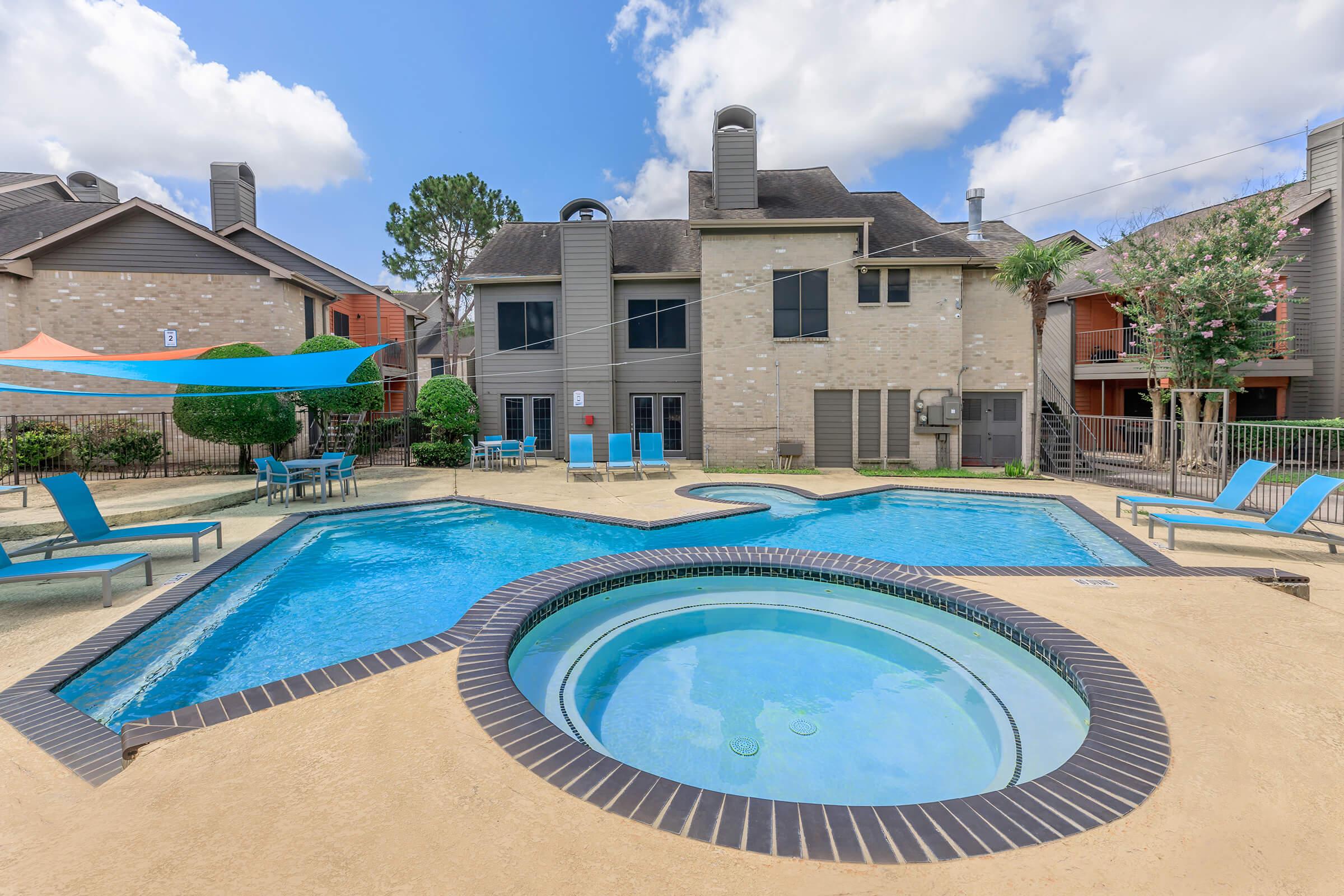 A spacious outdoor swimming area featuring two pools, one large and one smaller hot tub, surrounded by lounge chairs and shaded by colorful canopies. Modern apartment buildings are visible in the background with landscaped greenery and blue skies above.