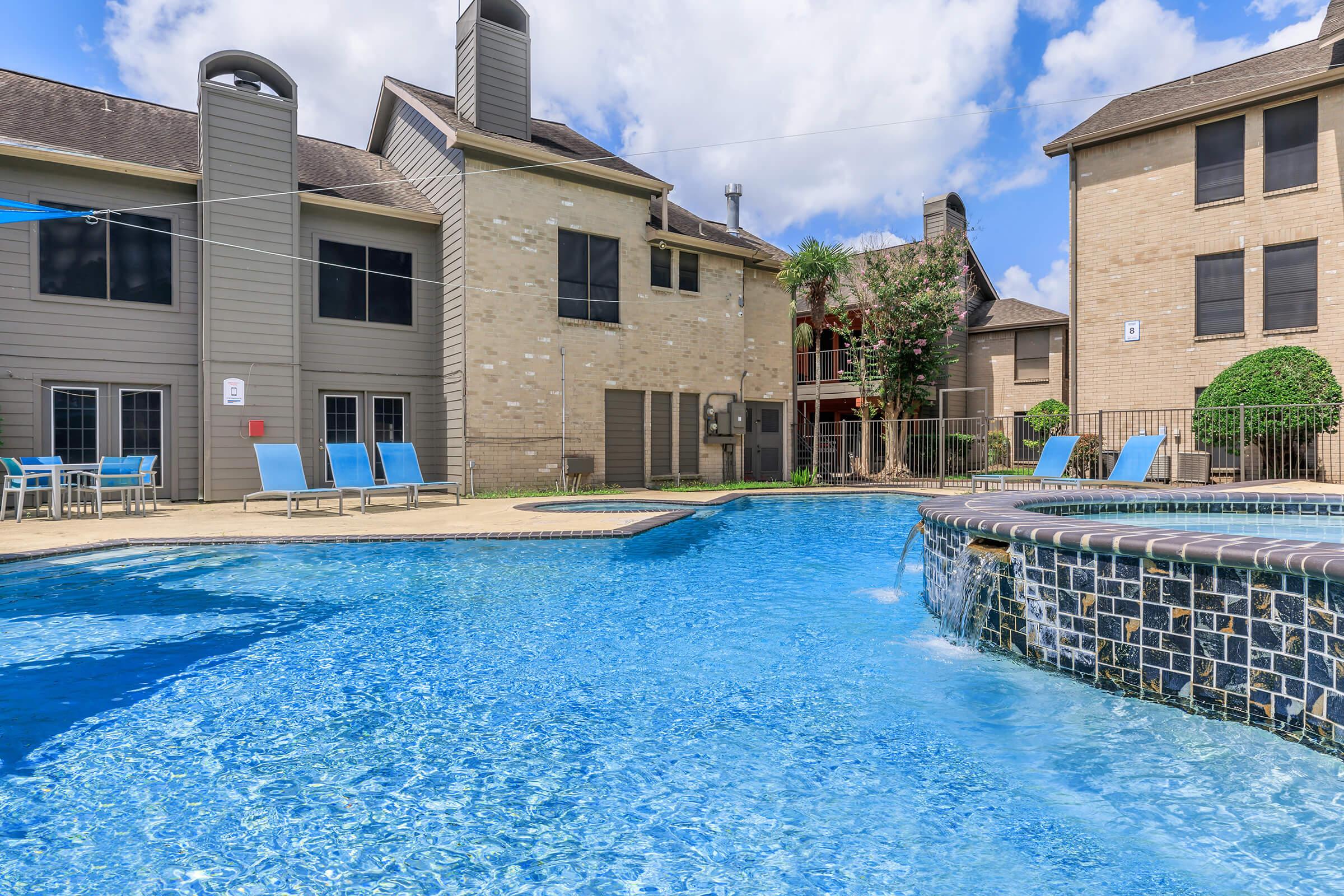 A clear blue swimming pool surrounded by lounge chairs and buildings with beige and gray exteriors. There are palm trees and a jacuzzi integrated into the pool area. The sky is partly cloudy, creating a vibrant and inviting atmosphere.