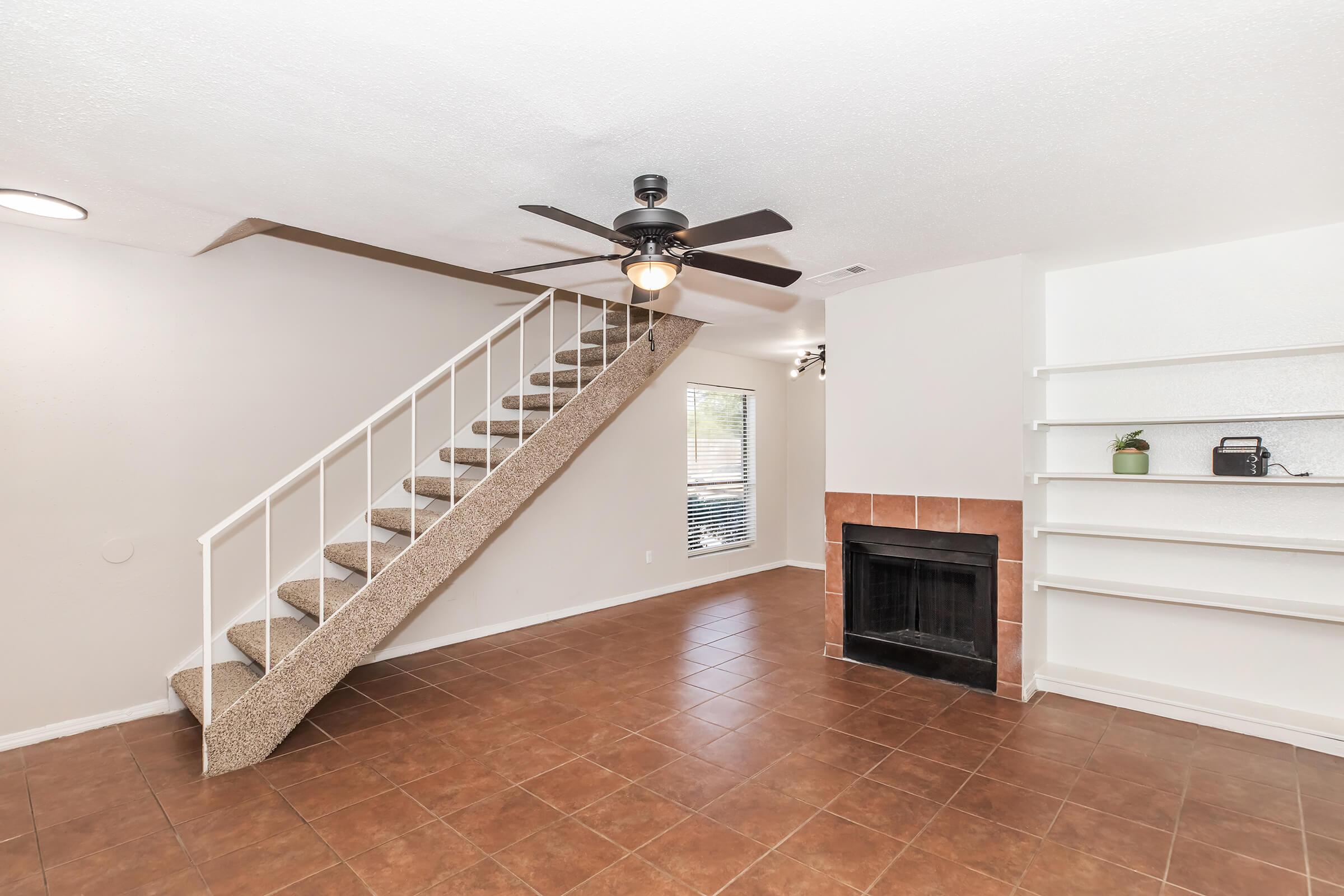 Interior of a living room featuring a staircase with carpeted steps, a ceiling fan, and a fireplace with a tiled surround. The space has light-colored walls and tiled flooring, with natural light coming through a window. There are built-in shelves on one wall.