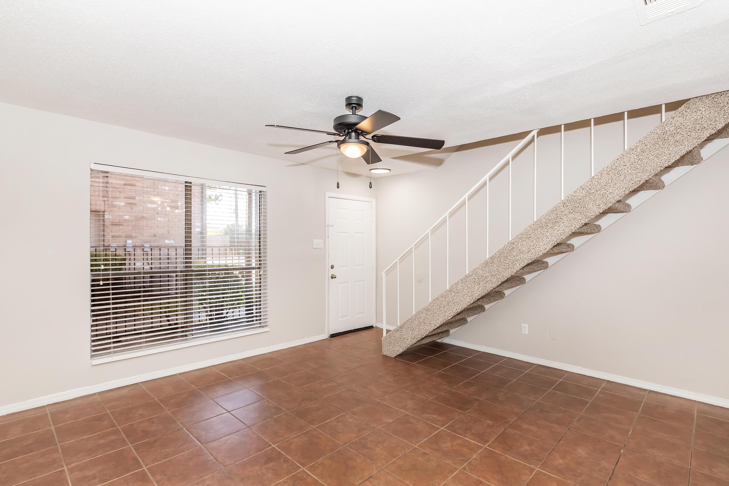 Interior view of a spacious living area featuring a ceiling fan, large window with blinds, tan tiled flooring, and a staircase with beige carpeting. The walls are painted in a light neutral color, and there is a door leading outside visible in the background.