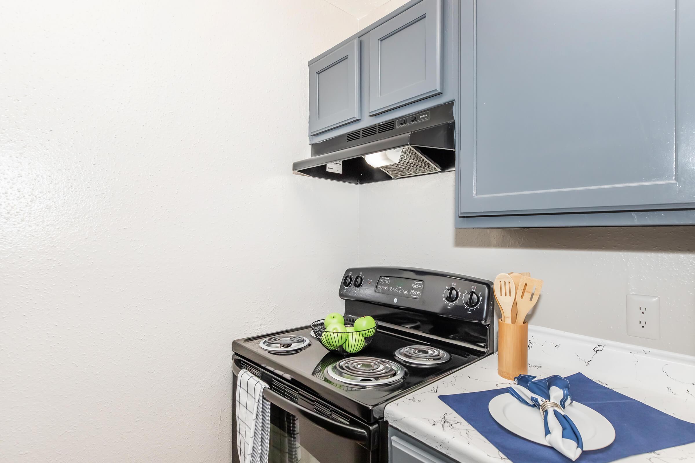 A modern kitchen with a black stove and oven, featuring a silver range hood above. Blue cabinets line the wall, and a white marble countertop holds a blue placemat with a white plate and utensils. A bowl of green apples adds a pop of color to the scene. The walls are plain and light-colored.