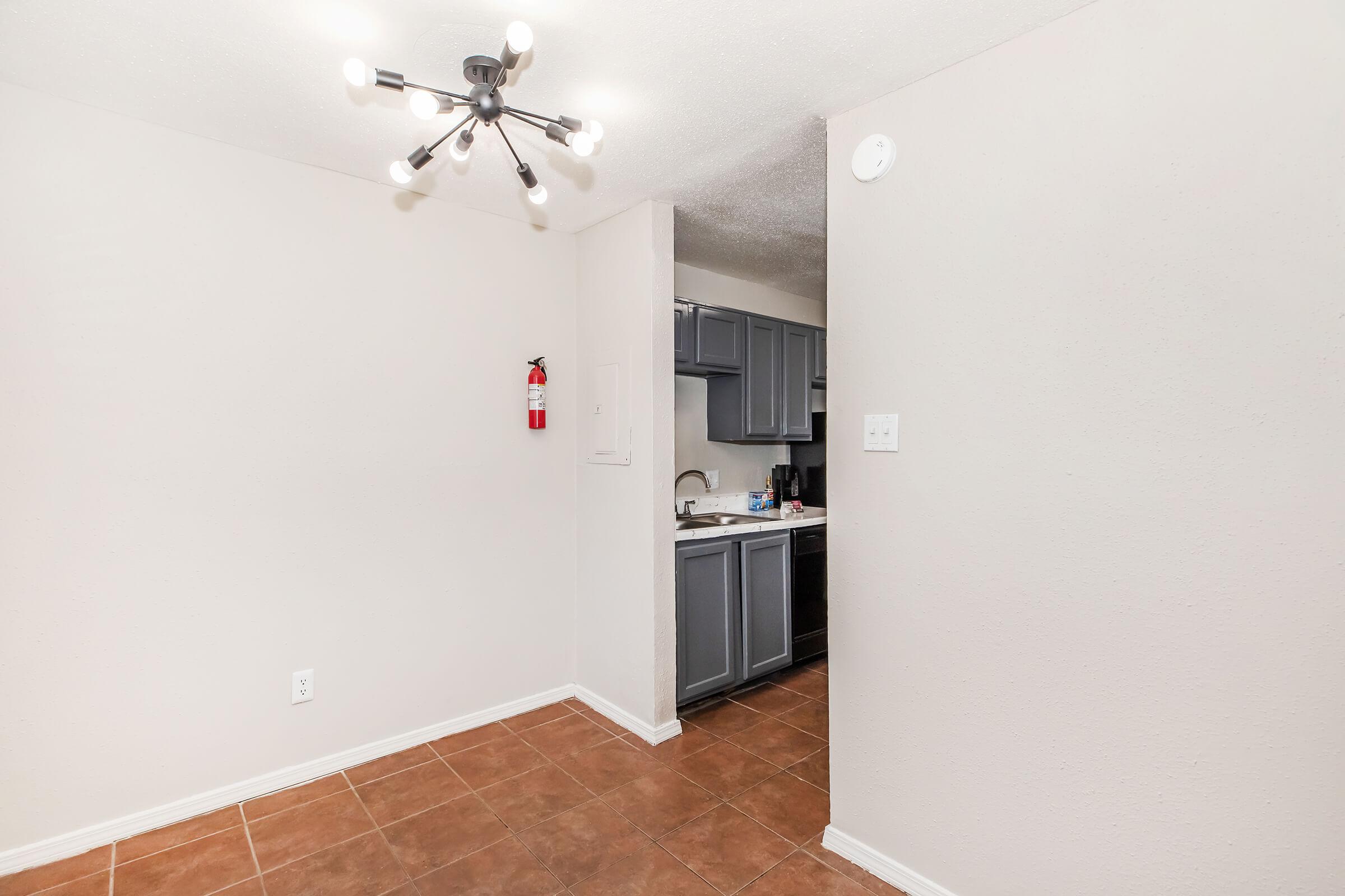 A view of a simple, well-lit room with beige walls and a tiled floor. A modern light fixture hangs from the ceiling. To the left, there is a fire extinguisher mounted on the wall. In the background, a kitchen area with dark cabinets can be seen. The space appears clean and minimalistic.